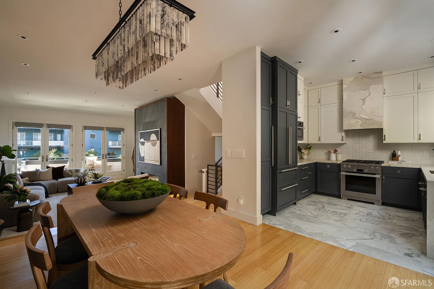 This open-concept living space features a dining area in the foreground with a large wooden table and a unique textured chandelier, transitioning into a modern kitchen with dark cabinetry and light stone countertops. A tall, dark stone-clad chimney breast separates the kitchen from the living area, which opens onto a balcony through large glass doors. The space is bright and airy, showcasing a blend of contemporary design elements and high-end finishes.