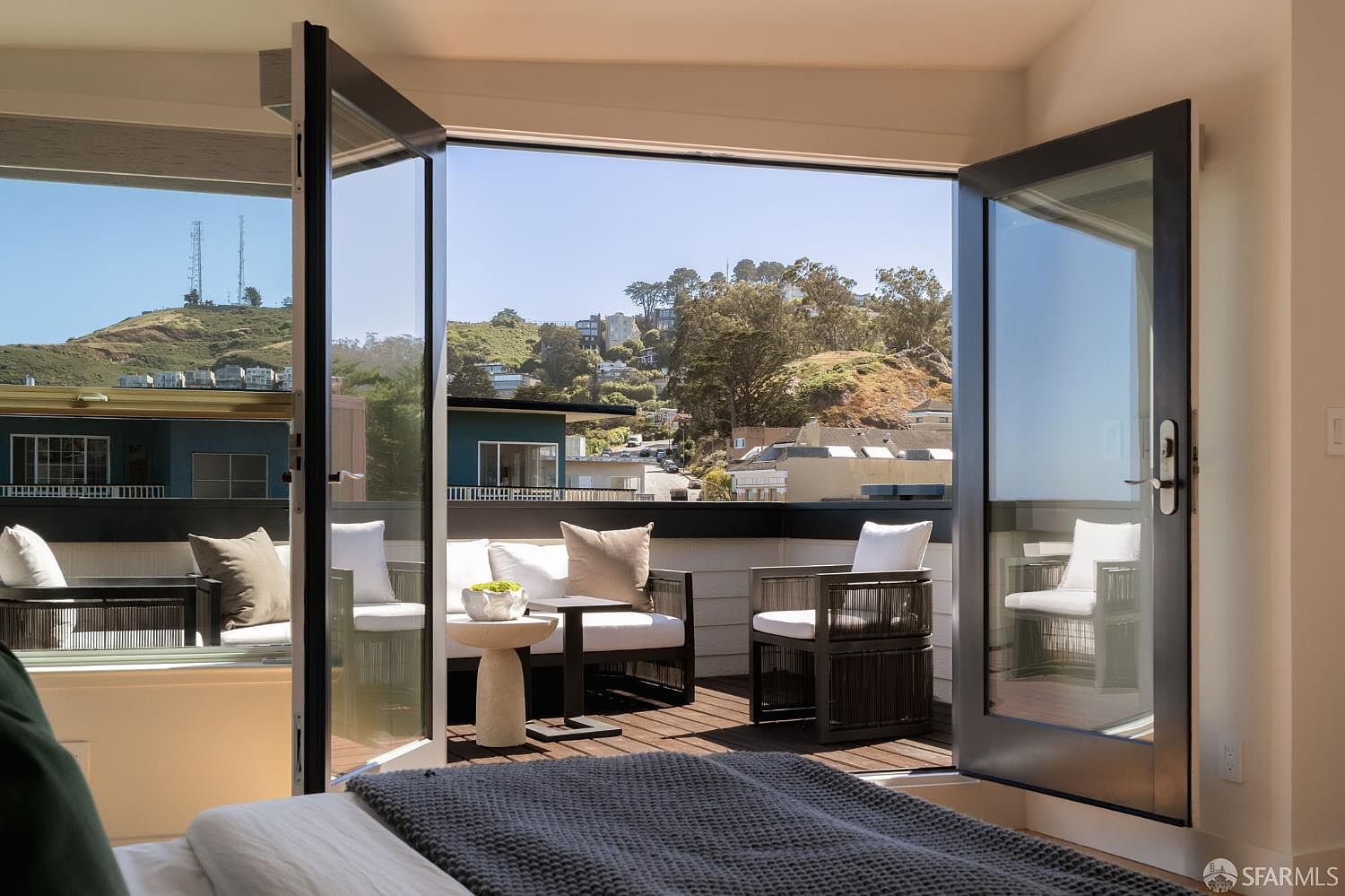 This primary bedroom features a seamless transition to a private outdoor deck through large, open glass doors. The deck is furnished with modern outdoor seating, including a sofa and armchairs, overlooking a scenic hillside neighborhood. The interior space is anchored by a bed with a textured gray throw, creating a serene and inviting atmosphere that emphasizes indoor-outdoor living.