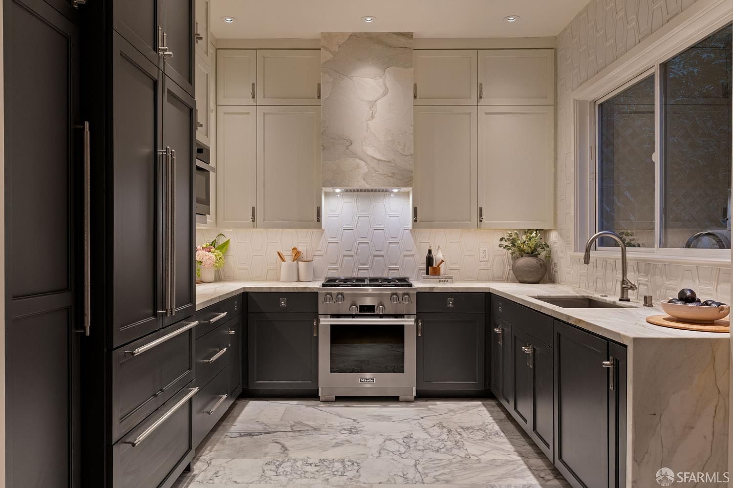 This elegant, U-shaped kitchen features a sophisticated contrast between dark charcoal lower cabinetry and light-toned upper cabinets. A striking stone-clad range hood serves as the focal point above a professional-grade stainless steel range, complemented by a geometric patterned backsplash and light-veined stone countertops. The space is finished with high-end appliances and polished stone flooring, creating a refined and modern culinary environment.