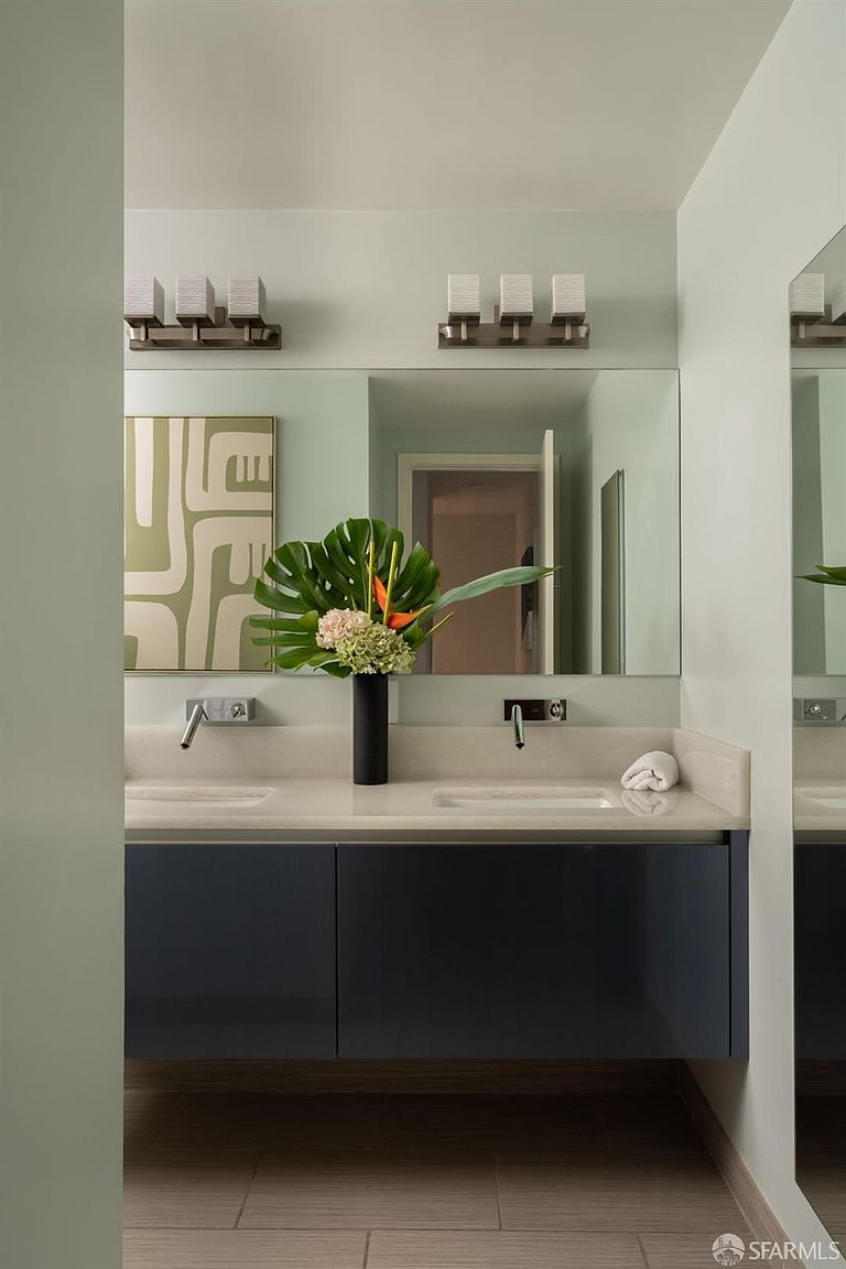 This modern primary bathroom features a sleek, wall-mounted vanity with dark cabinetry and a light-toned countertop, complemented by dual wall-mounted faucets. A large, frameless mirror spans the wall above the vanity, reflecting the room and a decorative piece of wall art, while two sets of three-light fixtures provide bright illumination. The space is accented by a central floral arrangement, creating a clean, sophisticated, and contemporary aesthetic.