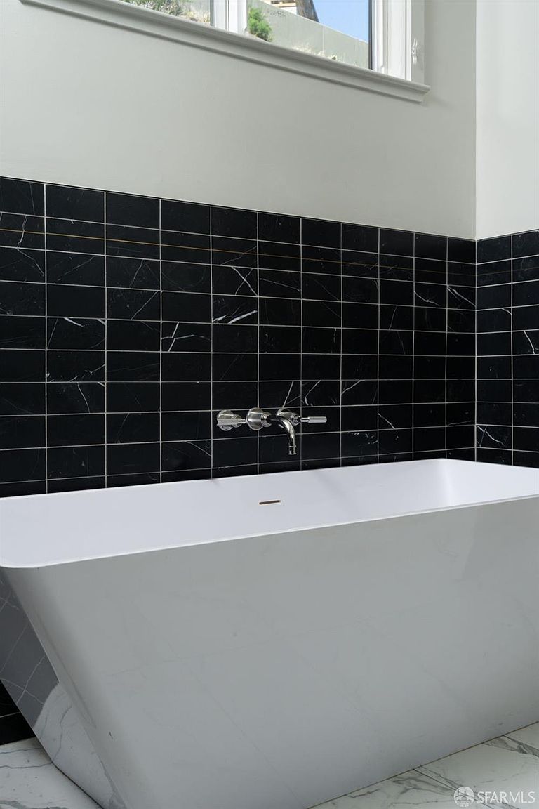 This modern bathroom features a sleek, white freestanding soaking tub set against a striking wall of black, rectangular stone tiles with white veining. A minimalist chrome wall-mounted faucet fixture is centered above the tub, providing a clean and contemporary aesthetic. The floor is finished in a coordinating white stone with subtle grey veining, creating a sophisticated and high-contrast look.