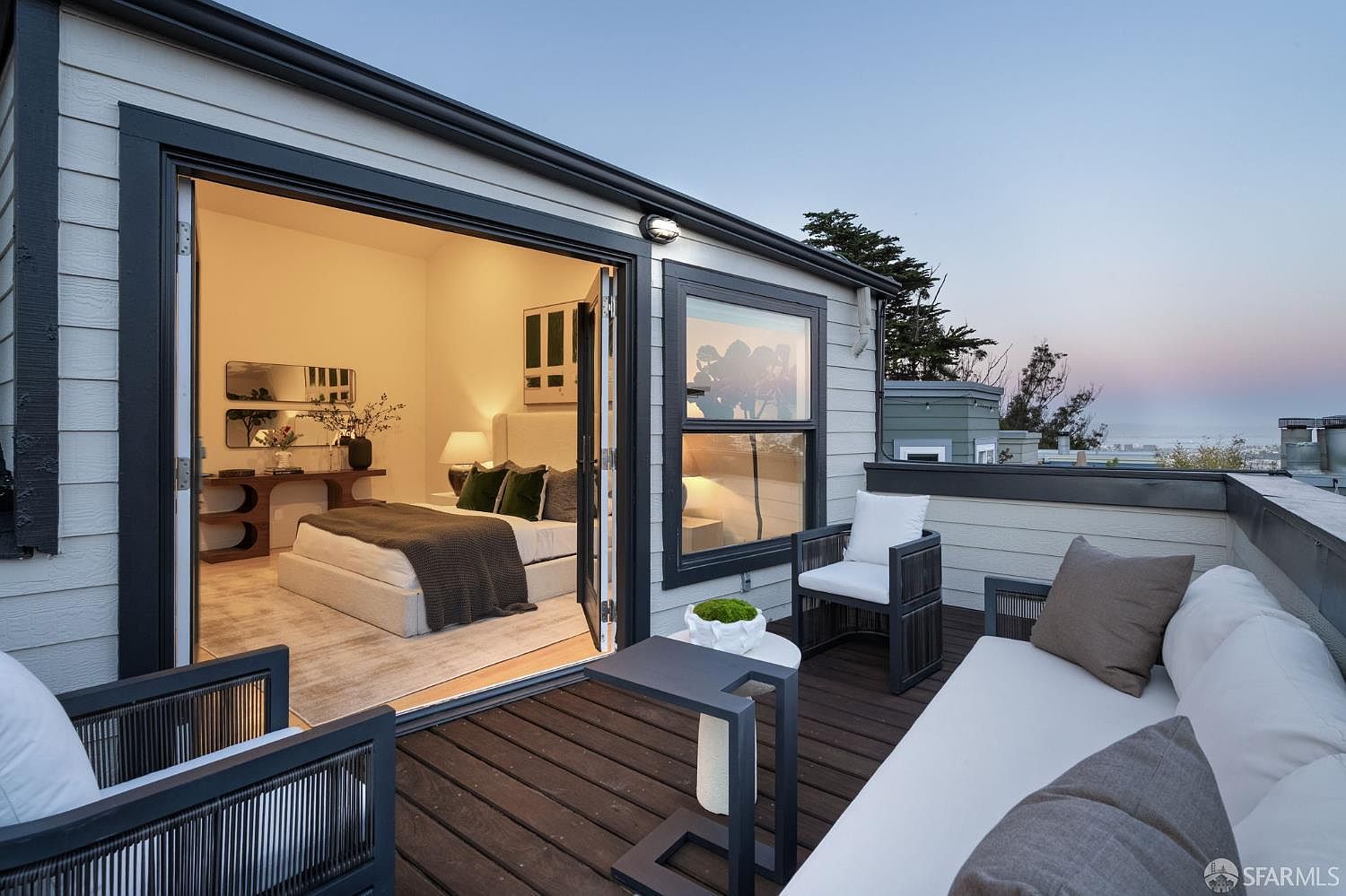 This image captures a modern, elevated deck featuring dark wood decking and comfortable outdoor lounge furniture, including a sofa and armchairs. Large glass doors open directly from the deck into a bright, neutrally decorated bedroom, creating a seamless indoor-outdoor living experience. The perspective looks out from the deck toward a distant horizon, highlighting the property's scenic vantage point.