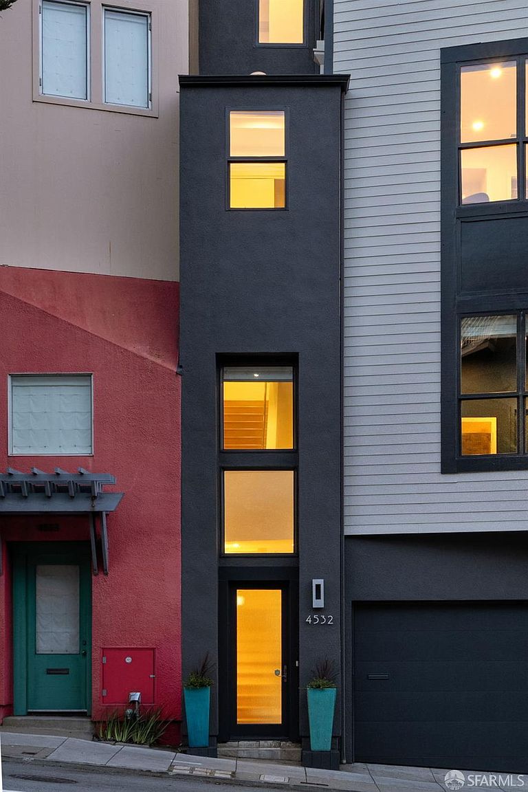 This narrow, multi-story townhouse features a striking modern facade with a dark, vertical central column that contrasts against the adjacent red and light-gray siding. The entrance is defined by a tall glass door and stacked windows that allow light to filter into the interior, while a sleek garage door sits to the right. The perspective is a straight-on street-level shot, emphasizing the unique, slender architectural profile of the home.