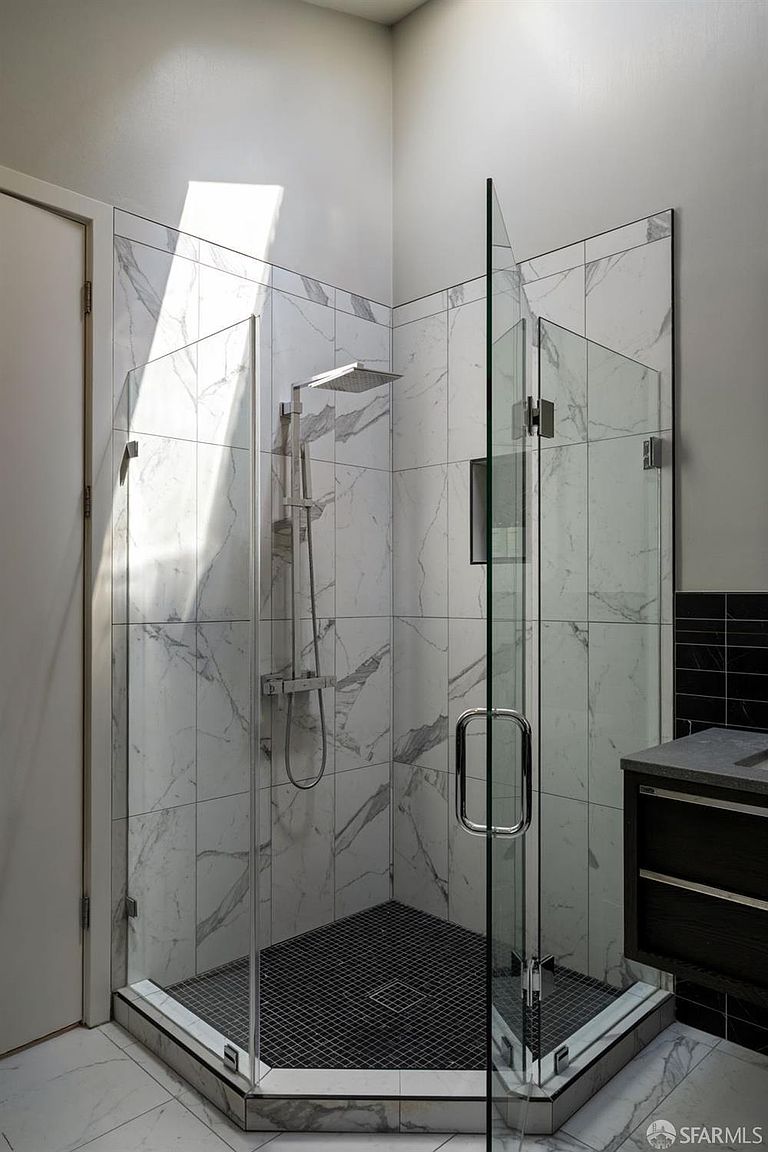 This modern walk-in shower features large-format light-colored stone tiles with subtle veining on the walls and a contrasting dark, small-format mosaic tile on the floor. A sleek, contemporary shower head is mounted on the wall, and the enclosure is defined by clear glass panels and a hinged door with a prominent metal handle. The perspective is eye-level, highlighting the clean lines and high-end finishes of the shower stall.
