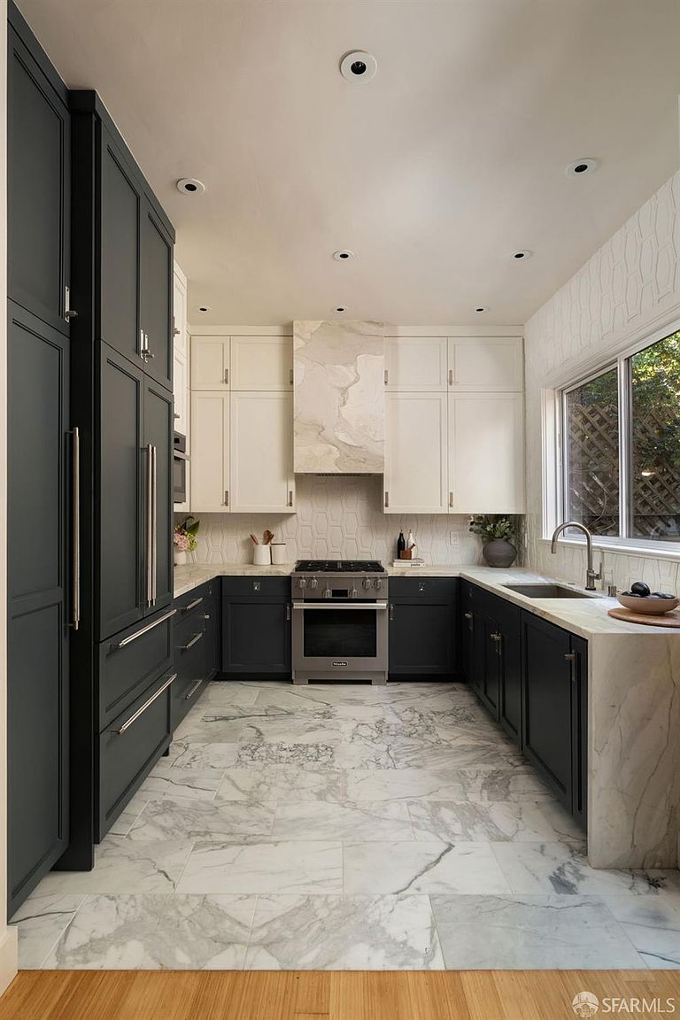 This modern kitchen features a striking two-tone color scheme with dark lower cabinets and tall pantry units contrasted by light upper cabinetry. A central range is framed by a custom stone-clad hood, while the space is finished with stone countertops and matching stone flooring. The perspective is a straight-on, symmetrical shot that highlights the clean lines and high-end finishes of the culinary space.