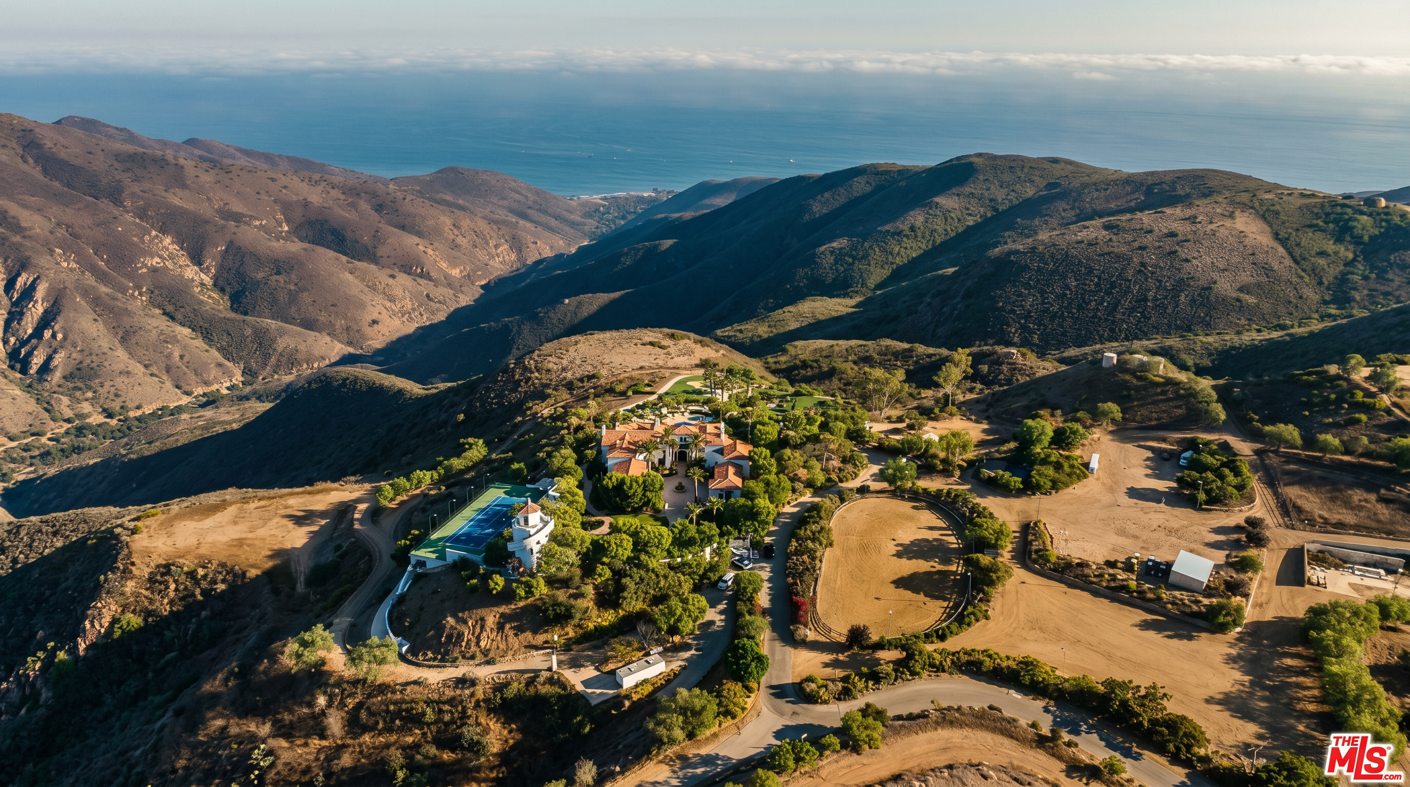 This stunning aerial view captures a sprawling estate nestled atop a rugged mountain ridge, offering panoramic vistas of the distant coastline and ocean. The property features a large main residence, a private tennis court, and an expansive equestrian riding ring, all surrounded by natural, arid landscape. The perspective emphasizes the secluded, luxurious nature of the estate, highlighting its unique integration with the dramatic topography.