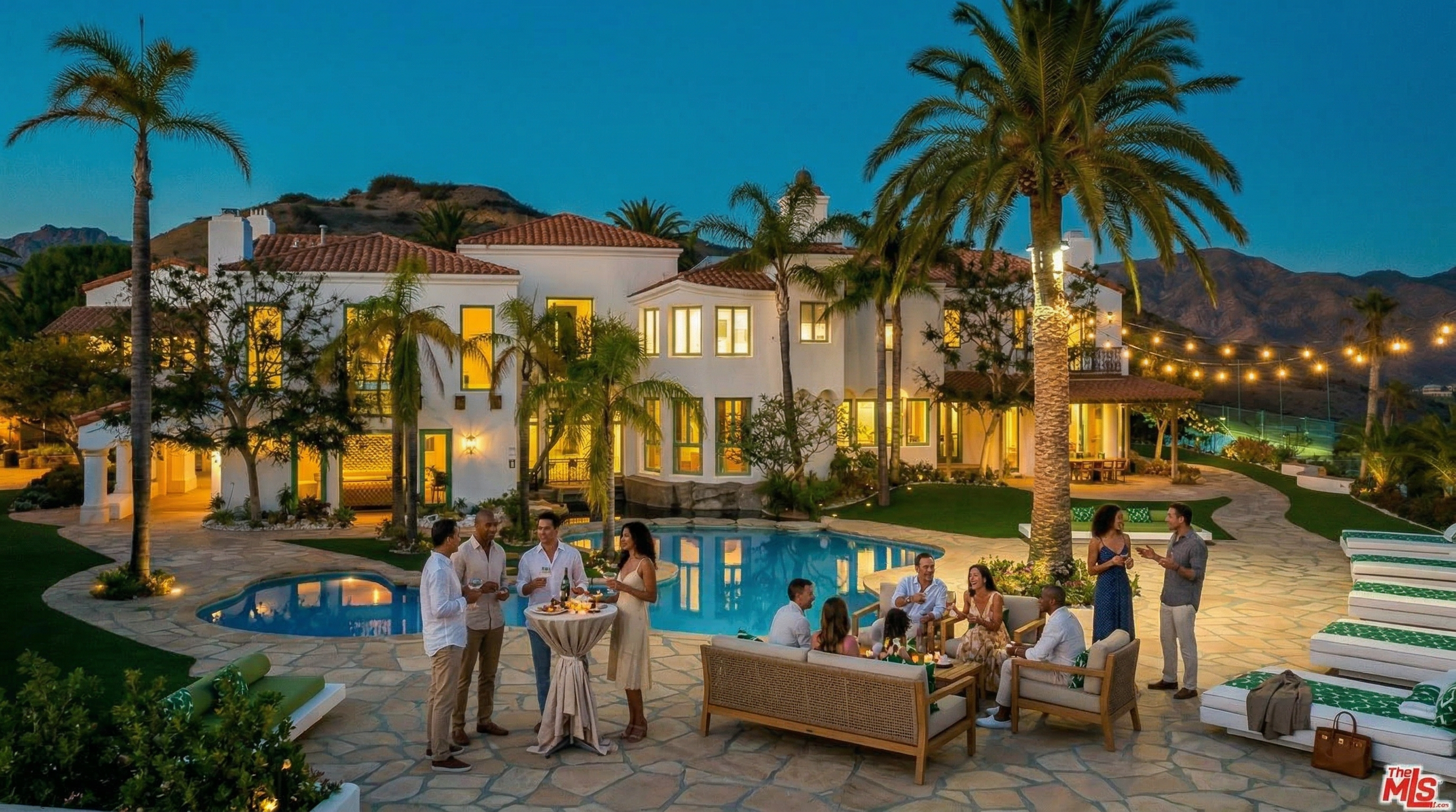 This stunning Mediterranean-style estate features a sprawling rear exterior with a custom-shaped swimming pool and expansive stone patio. The white stucco facade, accented by a red tile roof, is beautifully illuminated at dusk, creating a warm and inviting atmosphere against the backdrop of rugged mountains. The scene is completed by lush landscaping, mature palm trees, and a row of elegant lounge chairs, offering a luxurious resort-like feel.