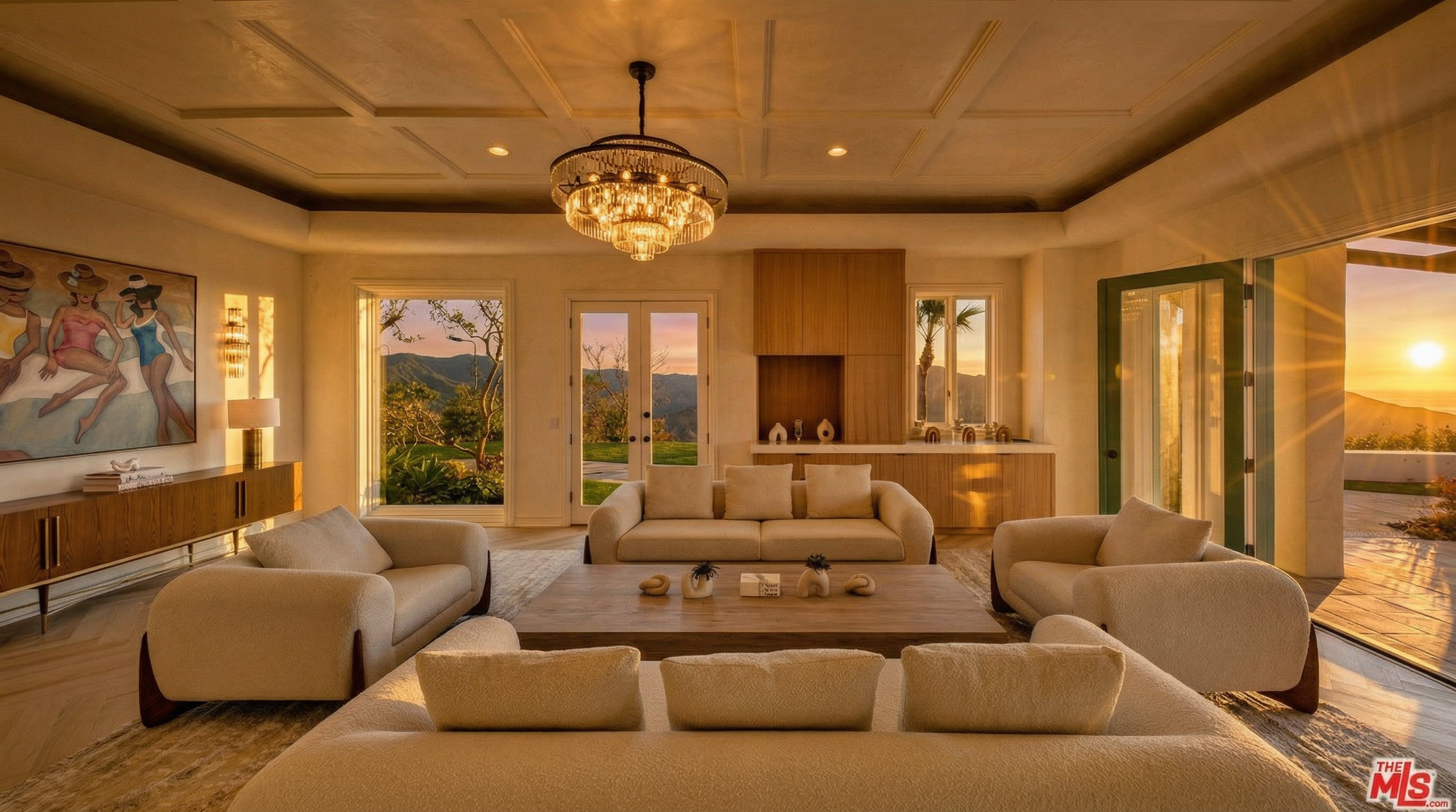 This elegant and spacious living room features a sophisticated neutral color palette with plush, modern cream-colored seating and a large central wooden coffee table. The coffered ceiling and ornate chandelier anchor the room, while expansive windows and open double doors frame breathtaking sunset views of the surrounding hills. A vibrant painting of swimmers adds a touch of artistic flair to this well-appointed, upscale living space.