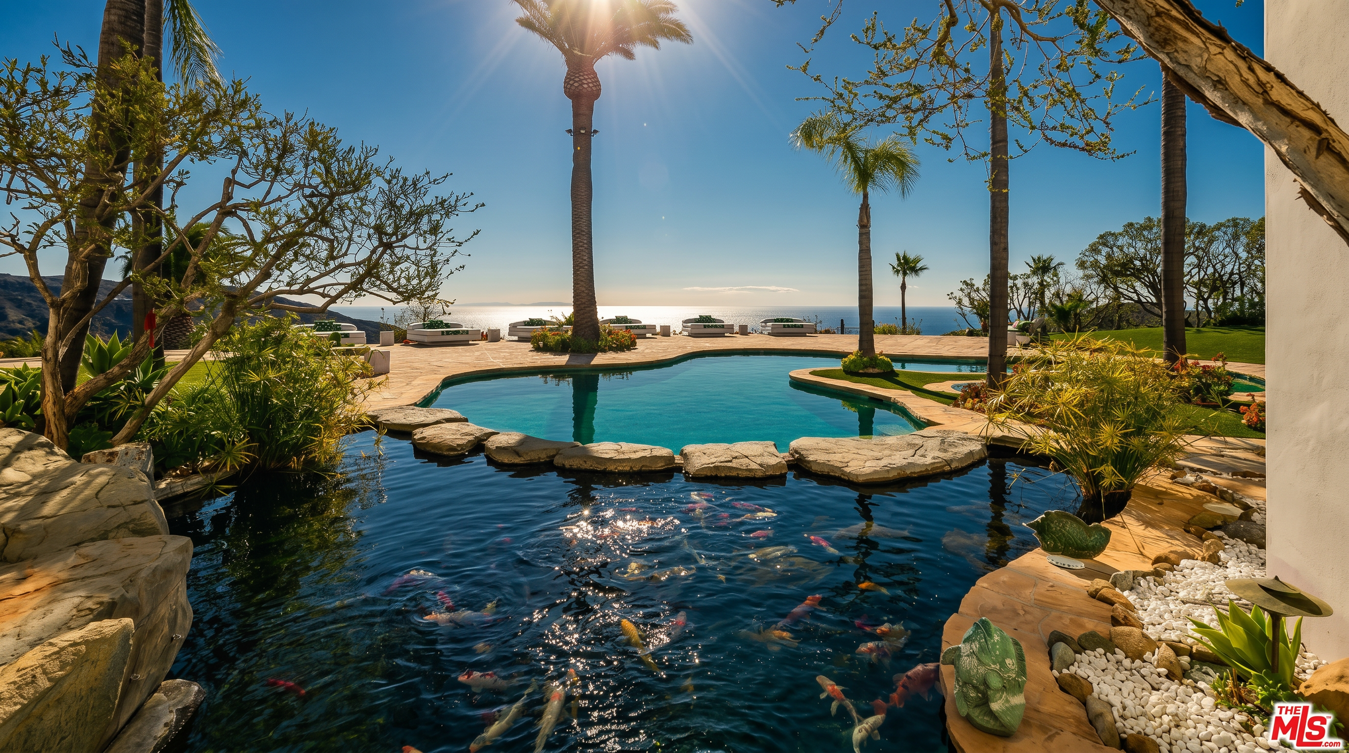 This stunning outdoor oasis features a large, free-form swimming pool seamlessly integrated with a foreground koi pond, separated by natural stone stepping stones. The expansive stone patio offers panoramic ocean views, framed by mature palm trees and lush landscaping, creating a serene and luxurious resort-like atmosphere. The perspective captures the tranquil water features in the foreground leading the eye toward the horizon, emphasizing the property's prime coastal location.