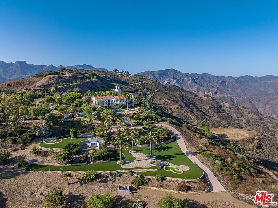 This expansive aerial view captures a luxurious Mediterranean-style estate perched atop a rugged hillside, surrounded by vast, arid mountain landscapes. The property features meticulously landscaped grounds, including a putting green, manicured lawns, and a winding driveway that leads up to the main residence. The scene conveys a sense of seclusion, grandeur, and high-end living in a dramatic natural setting.