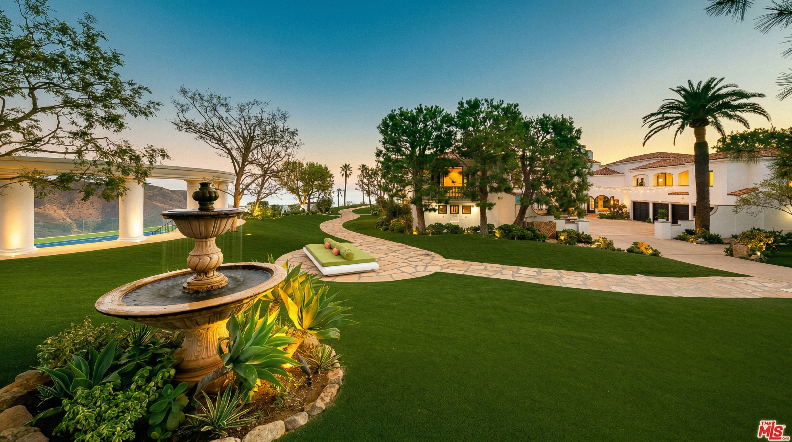 This expansive, meticulously manicured backyard features a classic tiered stone fountain as a focal point, surrounded by lush greenery and agave plants. A winding stone pathway leads through the vibrant green lawn toward a luxurious Mediterranean-style estate, complete with a palm tree and a serene outdoor lounge area. The scene is captured from a low-angle perspective, emphasizing the vastness of the property and the peaceful, high-end atmosphere of the landscape.