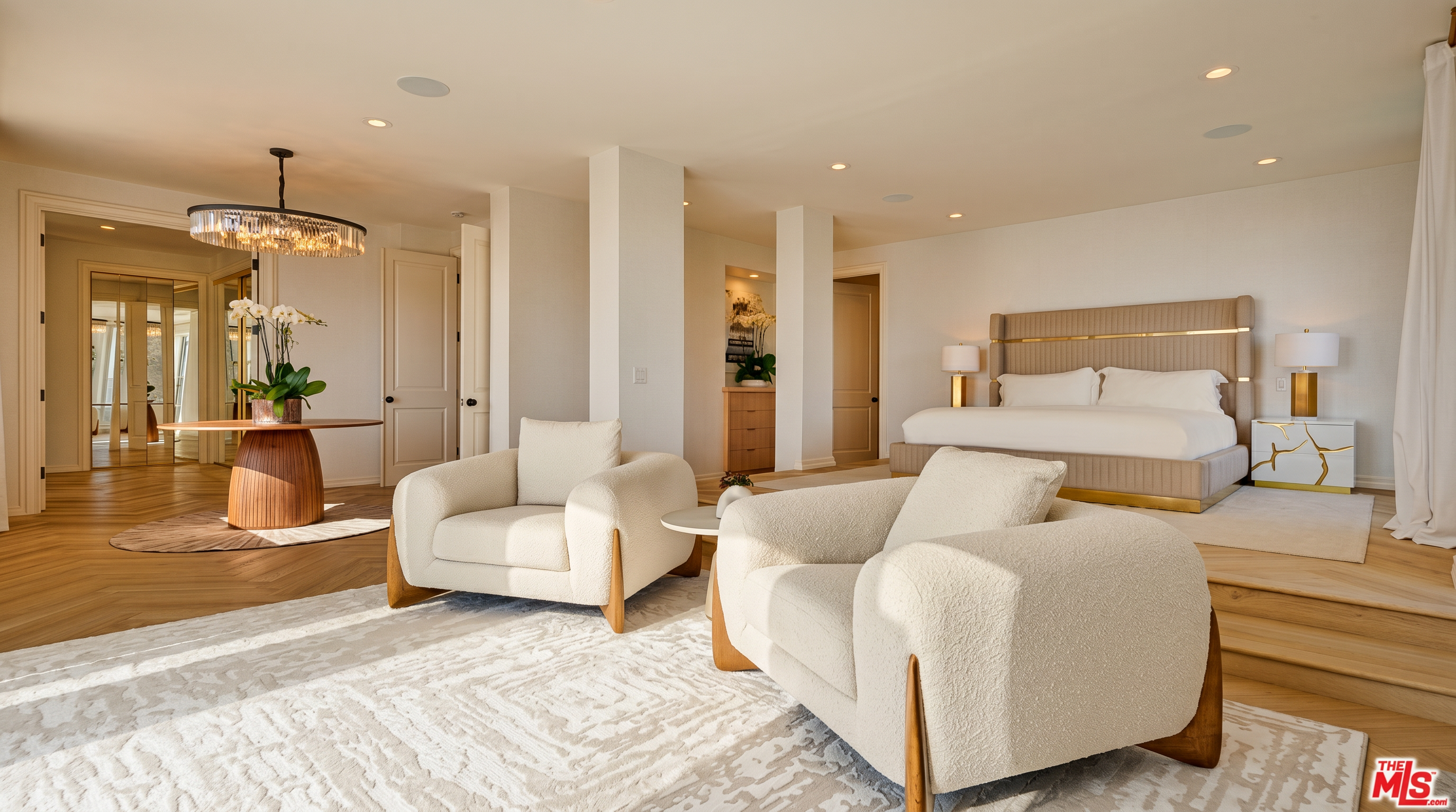 This expansive primary bedroom suite features a sophisticated, neutral color palette with a raised platform area for the bed and a cozy seating area in the foreground. The room is accented by elegant modern furniture, including two plush white armchairs, a statement chandelier, and a sleek, upholstered headboard. The open-concept layout, combined with light wood flooring and recessed lighting, creates a bright, airy, and luxurious atmosphere.