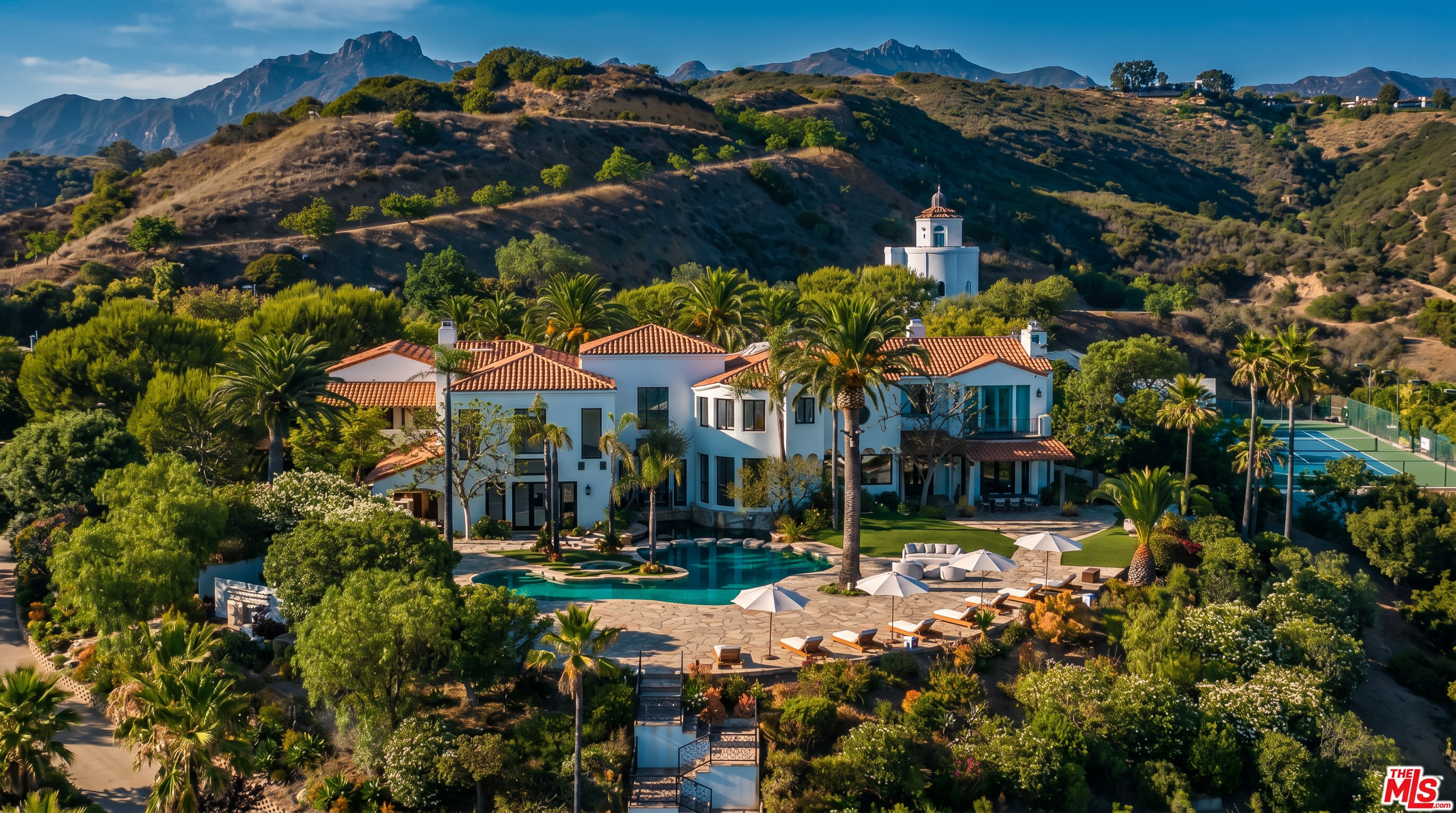 This stunning aerial view captures a sprawling Mediterranean-style estate nestled against a backdrop of rugged mountains. The property features a large, custom-shaped swimming pool, expansive landscaped grounds with mature palm trees, and a distinctive white tower structure on the hillside. The architecture showcases classic terracotta roof tiles and white stucco walls, creating a luxurious and serene atmosphere.