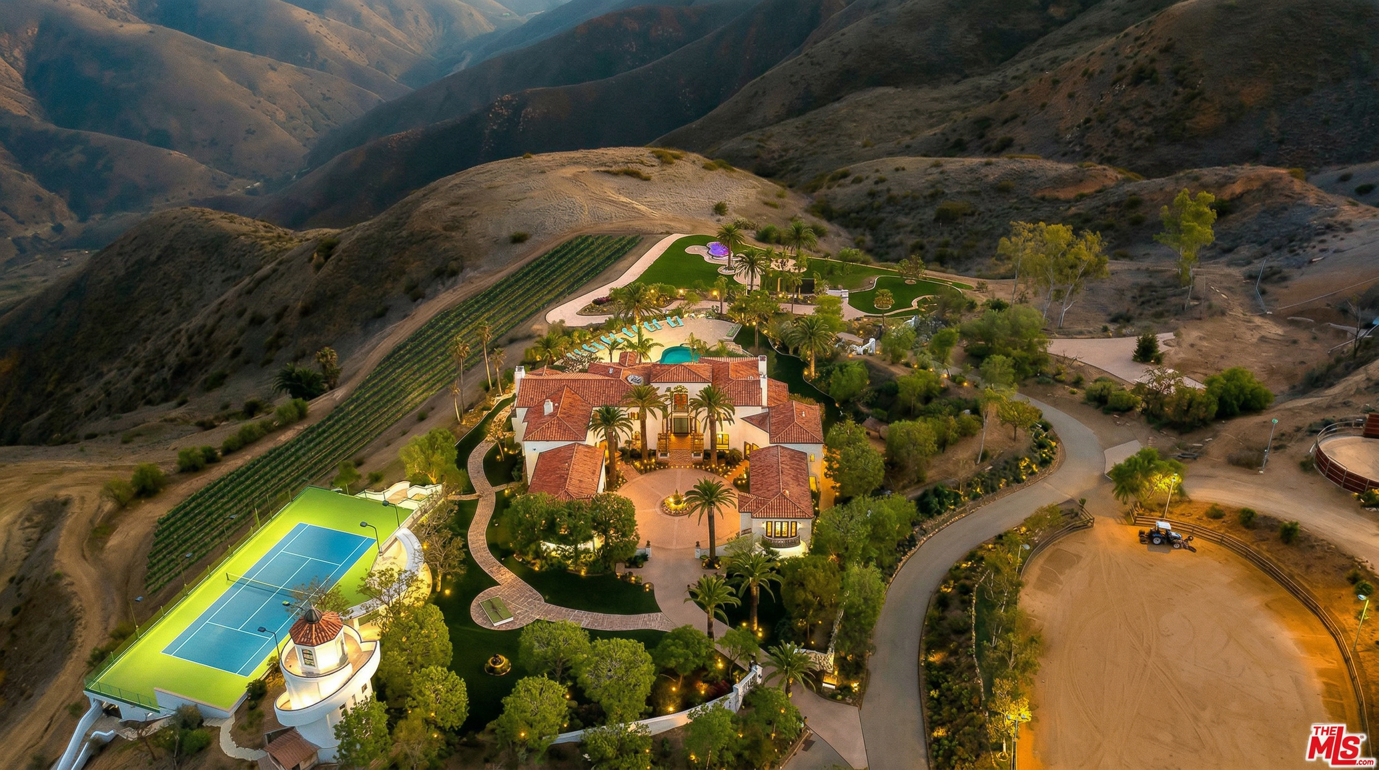 This stunning aerial perspective captures a sprawling Mediterranean-style estate nestled within a vast, hilly landscape. The property features a grand residence with a terracotta tile roof, a private tennis court, a swimming pool, and extensive manicured grounds including a vineyard. The cinematic high-angle shot emphasizes the luxury and seclusion of the estate, showcasing its integration into the natural topography.
