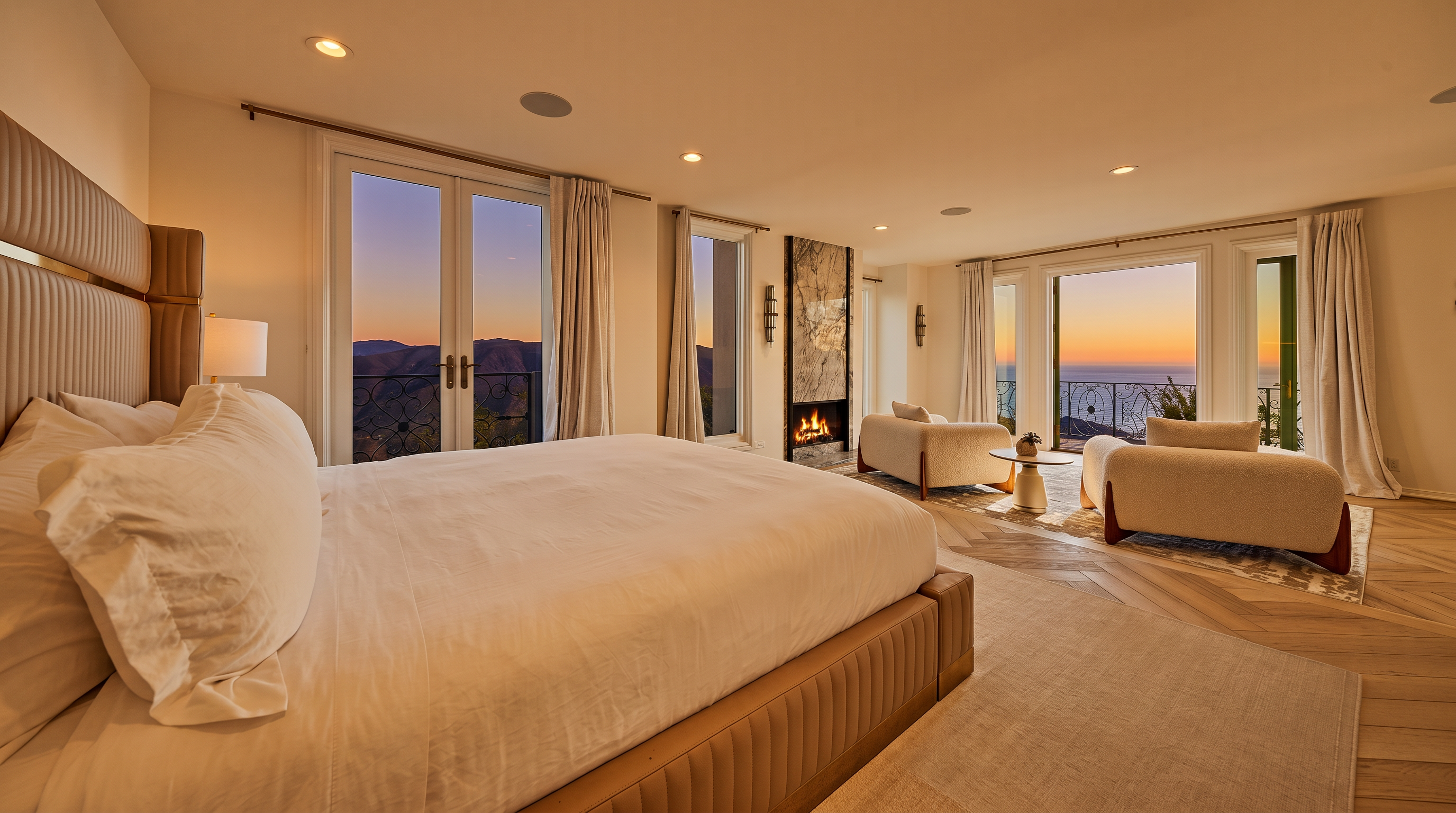 This luxurious primary bedroom features a spacious layout with a large bed, a cozy seating area, and a modern fireplace. The room is bathed in natural light from multiple glass doors that open to a balcony with stunning ocean and mountain views. The neutral color palette, herringbone wood flooring, and elegant furnishings create a serene and sophisticated atmosphere.