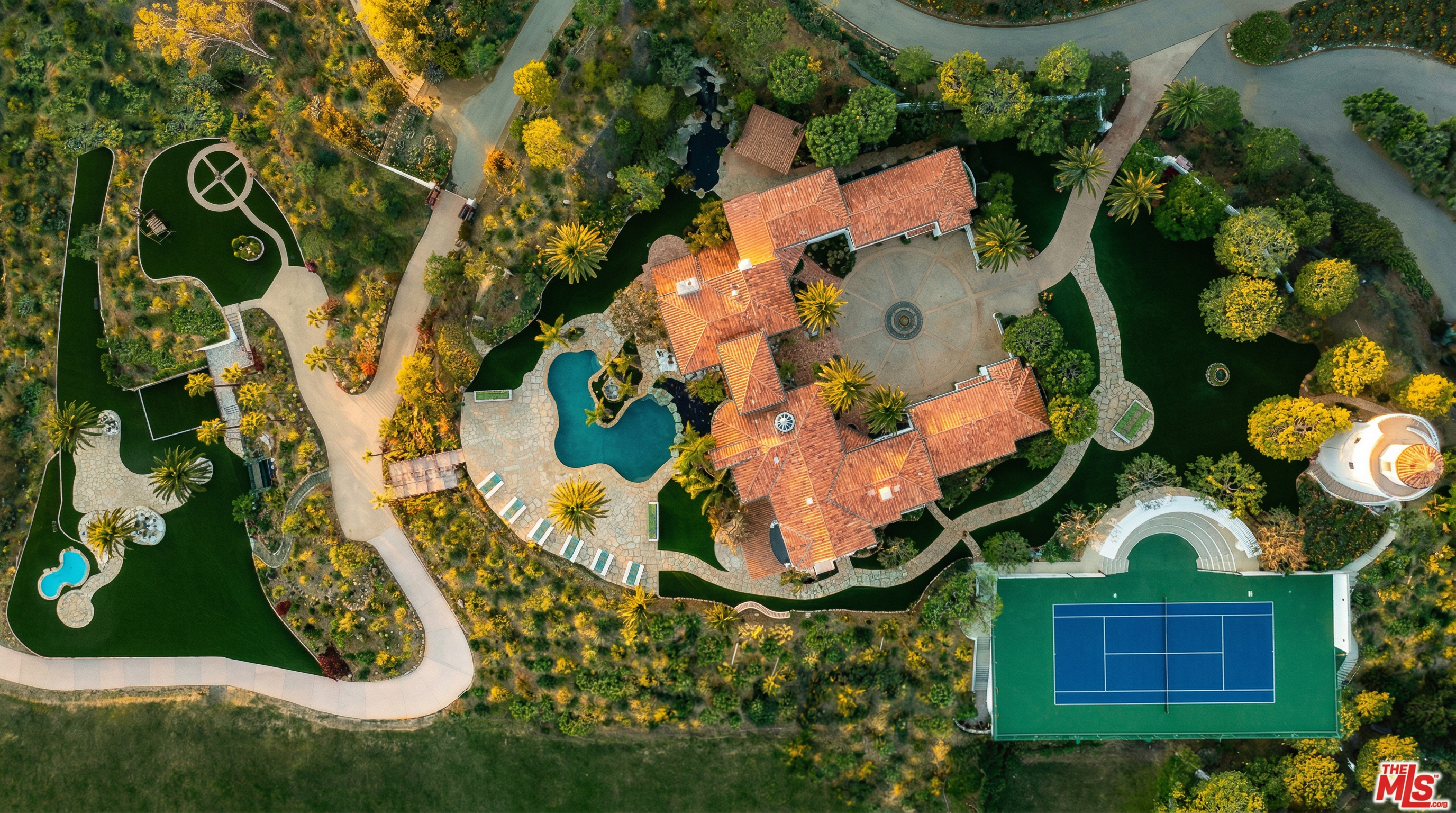 This stunning aerial perspective captures a sprawling Mediterranean-style estate nestled within a lush, landscaped property. The expansive grounds feature a large central courtyard, a custom-shaped swimming pool, a private tennis court, and winding pathways that weave through manicured gardens and mature trees. The high-angle shot emphasizes the grandeur and privacy of the residence, showcasing its intricate roofline and extensive outdoor recreational amenities.