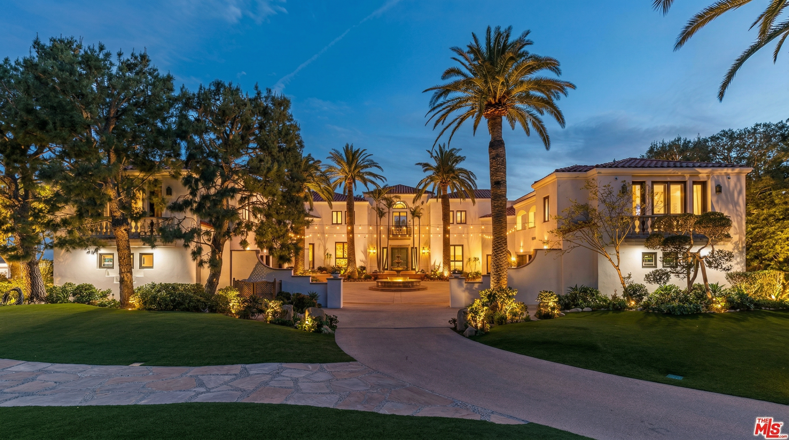 This stunning Mediterranean-style estate is captured at twilight, showcasing its grand symmetrical architecture and warm, inviting exterior lighting. A sweeping stone driveway leads to a central fountain, framed by lush green lawns and mature palm trees that enhance the property's luxurious curb appeal. The soft glow of string lights and interior illumination creates a sophisticated and welcoming atmosphere for this impressive residence.