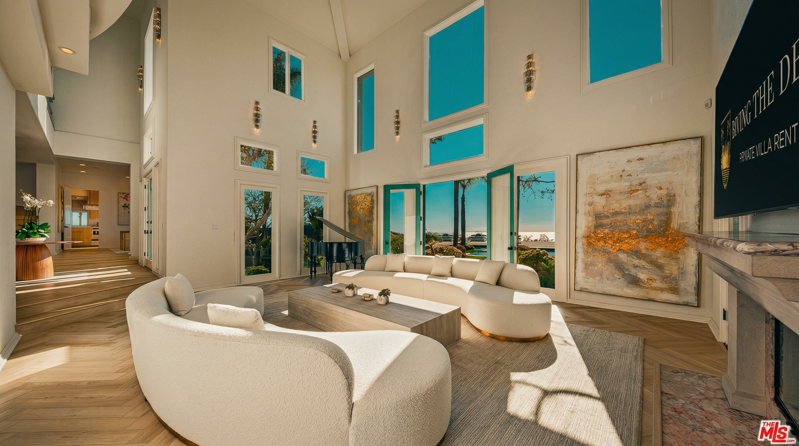 This expansive, double-height living room exudes luxury with its bright, airy atmosphere, featuring a curved white sofa set, a large wooden coffee table, and a grand piano. The space is anchored by a striking marble fireplace and a large abstract painting, while floor-to-ceiling windows and glass doors provide a seamless connection to the outdoor scenery. The light-toned herringbone flooring and minimalist decor create a sophisticated, high-end aesthetic perfect for elegant entertaining.
