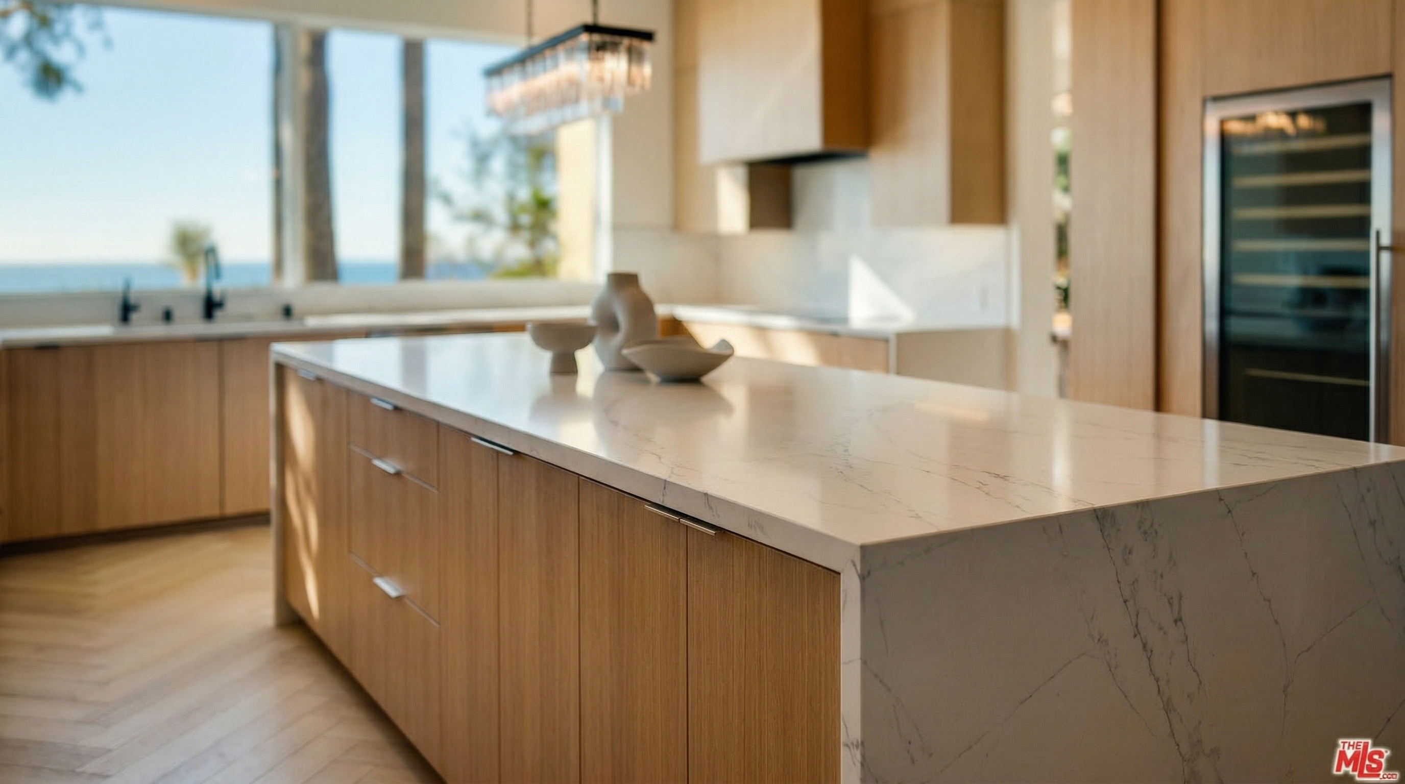 This sleek, modern kitchen features natural wood cabinetry and a large waterfall-edge island topped with veined white quartz. A glass pendant light fixture adds a touch of elegance, while the wide window offers stunning views of the ocean. The minimalist aesthetic and high-end finishes create a bright, airy, and sophisticated atmosphere.