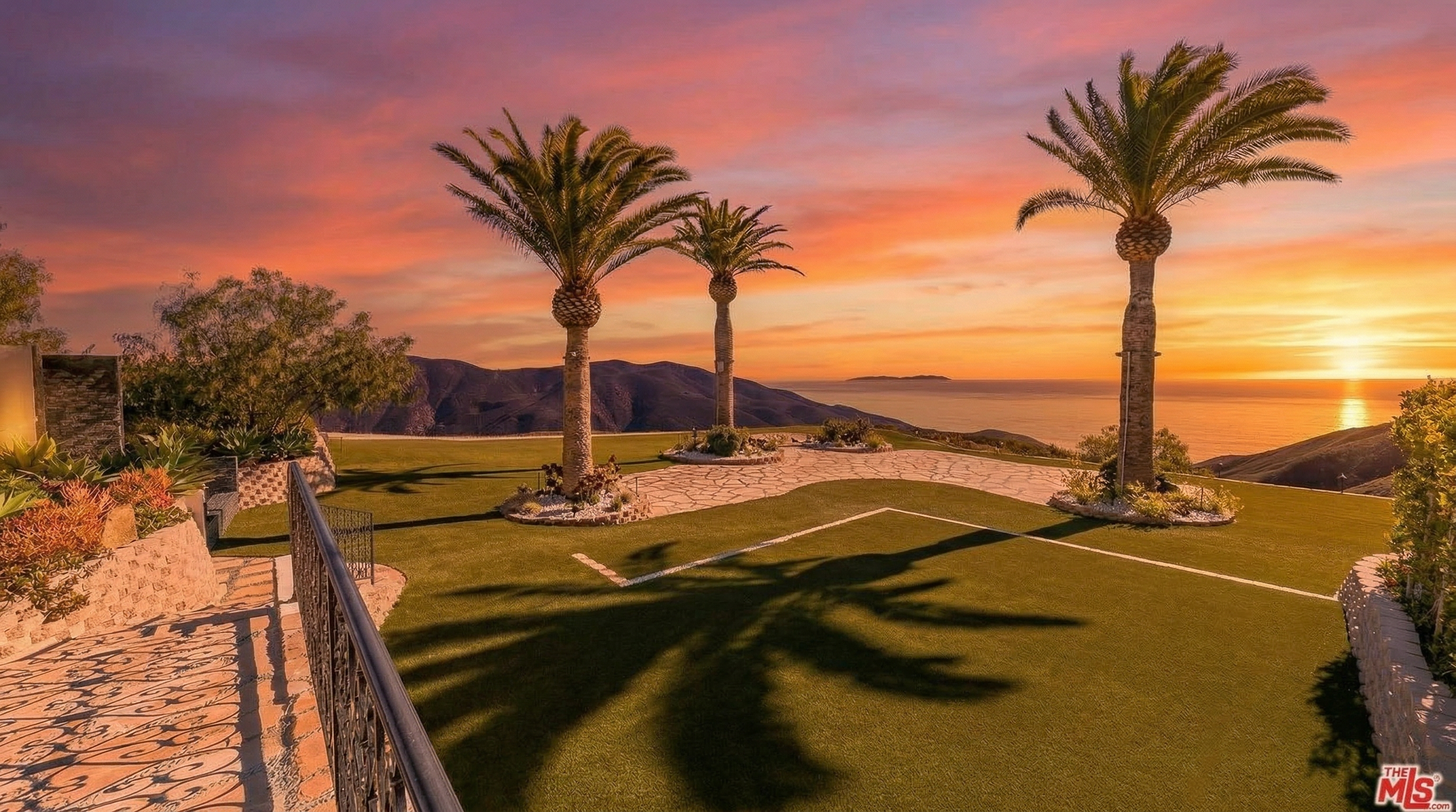This breathtaking backyard features a pristine manicured lawn and stone-paved pathways framed by three elegant palm trees. Set against a dramatic sunset backdrop overlooking the ocean and rolling hills, the space creates an inviting and luxurious outdoor retreat. The meticulous landscaping and sweeping coastal views suggest a high-end property perfect for outdoor relaxation or entertaining.