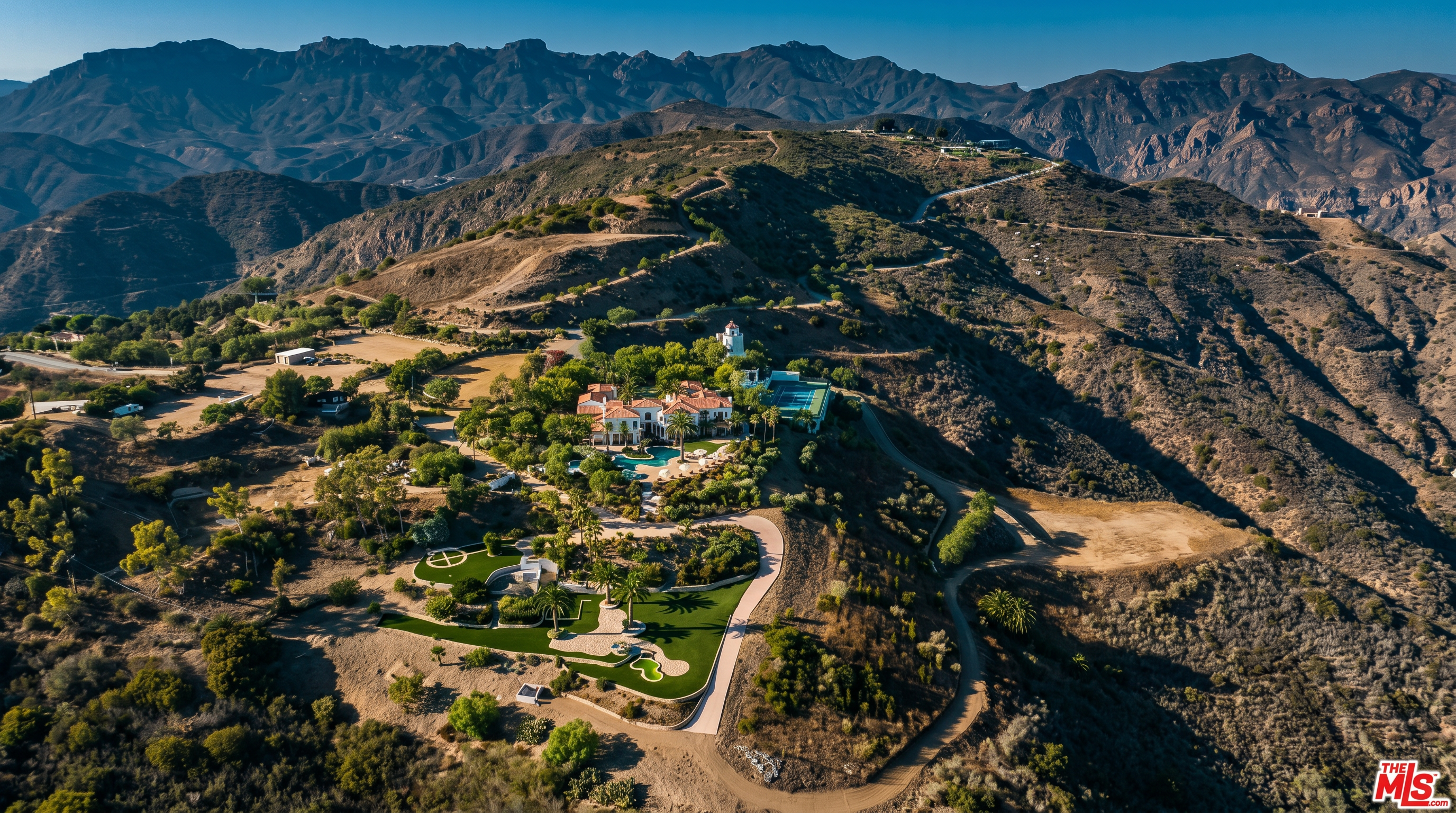 This stunning aerial perspective captures a sprawling Mediterranean-style estate nestled high within a rugged, mountainous landscape. The property features a large main residence with a terracotta roof, a sparkling swimming pool, a private tennis court, and meticulously landscaped grounds that include a putting green. The vast, elevated setting offers a sense of seclusion and grandeur, highlighting the seamless integration of luxury living with the natural, arid beauty of the surrounding hills.