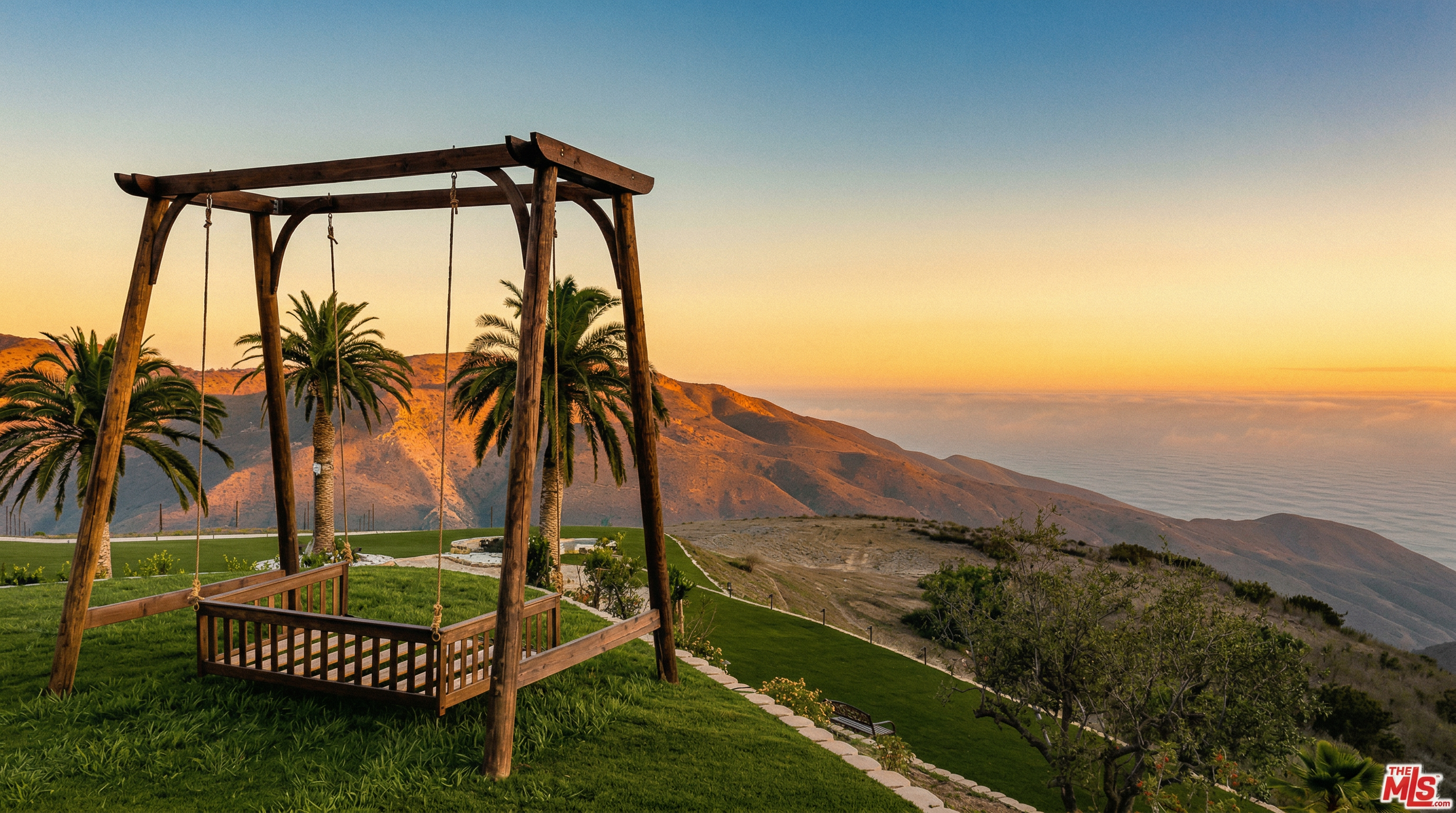 This stunning outdoor space features a large, wooden swing set positioned on a manicured lawn, offering breathtaking panoramic views of rolling hills and a distant cloud-covered horizon at sunset. The scene is framed by elegant palm trees and a stone-bordered path, creating a serene and luxurious atmosphere. The perspective captures the vastness of the landscape, emphasizing the property's prime location and tranquil outdoor living potential.