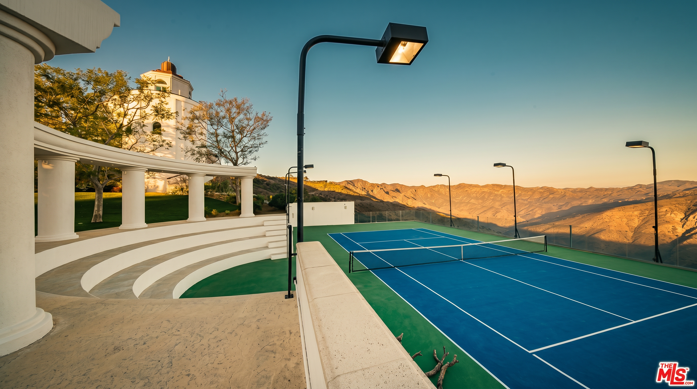 This image showcases a private tennis court set against a dramatic backdrop of rolling hills and a clear blue sky. The court features a vibrant blue and green surface, complemented by a curved, white colonnade and tiered seating area that adds an elegant, architectural touch. The perspective is elevated, emphasizing the expansive outdoor space and the luxurious, resort-like atmosphere of the property.