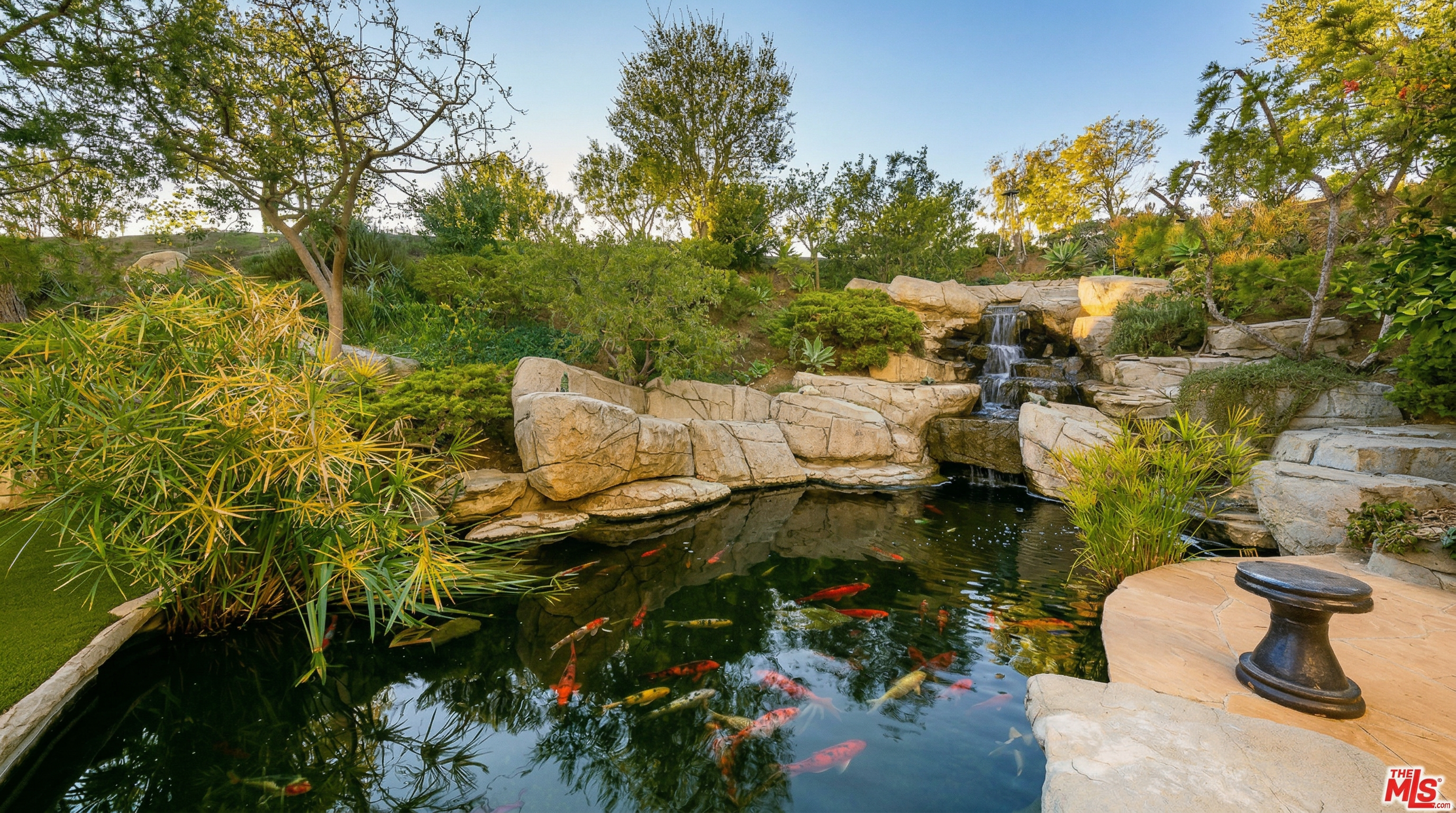 This serene backyard oasis features a naturalistic koi pond complete with a cascading rock waterfall and lush, landscaped surroundings. The scene is framed by mature trees and decorative boulders, creating a tranquil and private atmosphere. A small, dark pedestal table sits on the stone patio edge, providing a perfect vantage point to enjoy the peaceful water feature.