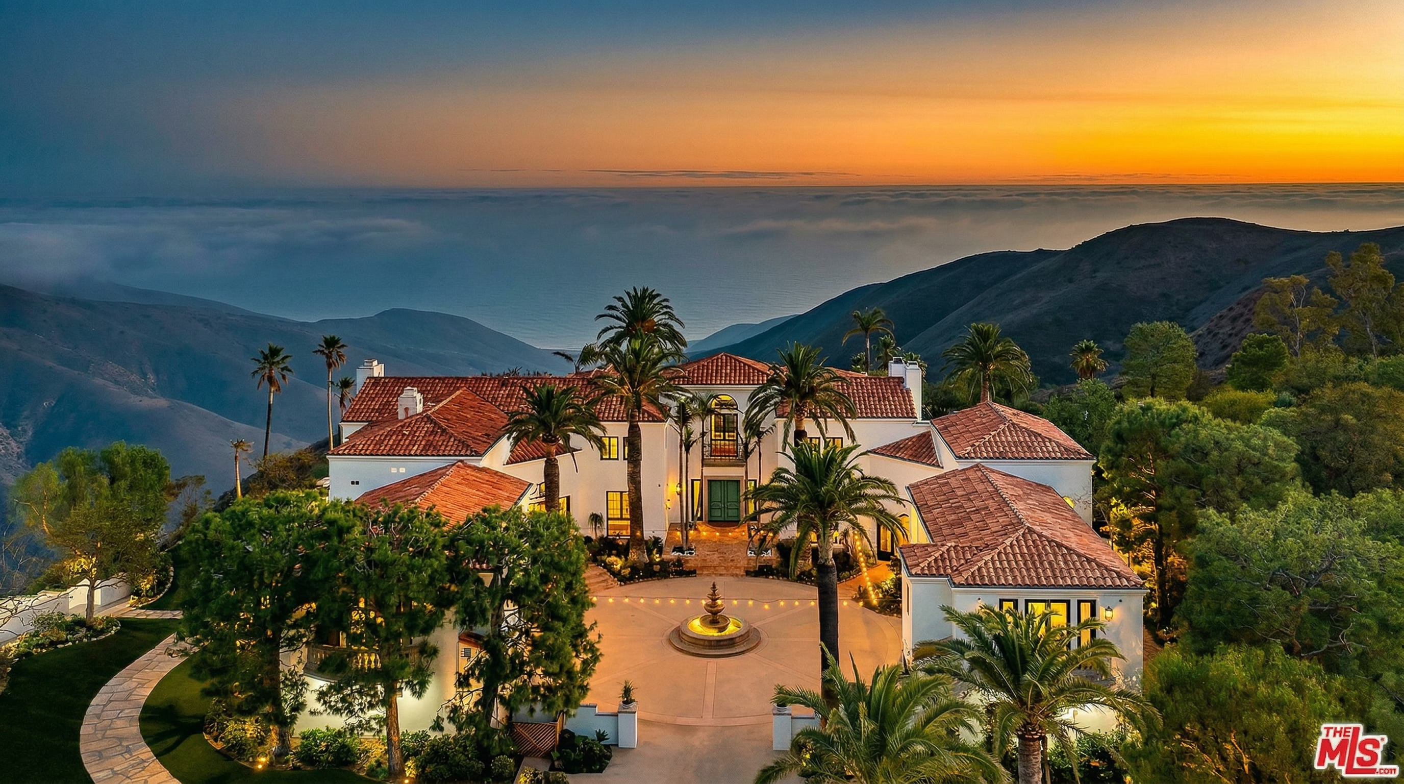 This stunning aerial view captures a luxurious Mediterranean-style estate perched atop a mountain ridge at sunset. The property features a grand circular driveway with a central fountain, terracotta-tiled roofs, and lush palm trees, all set against a dramatic backdrop of rolling hills and a sea of clouds. The perspective emphasizes the home's secluded, high-end location and its impressive architectural scale.
