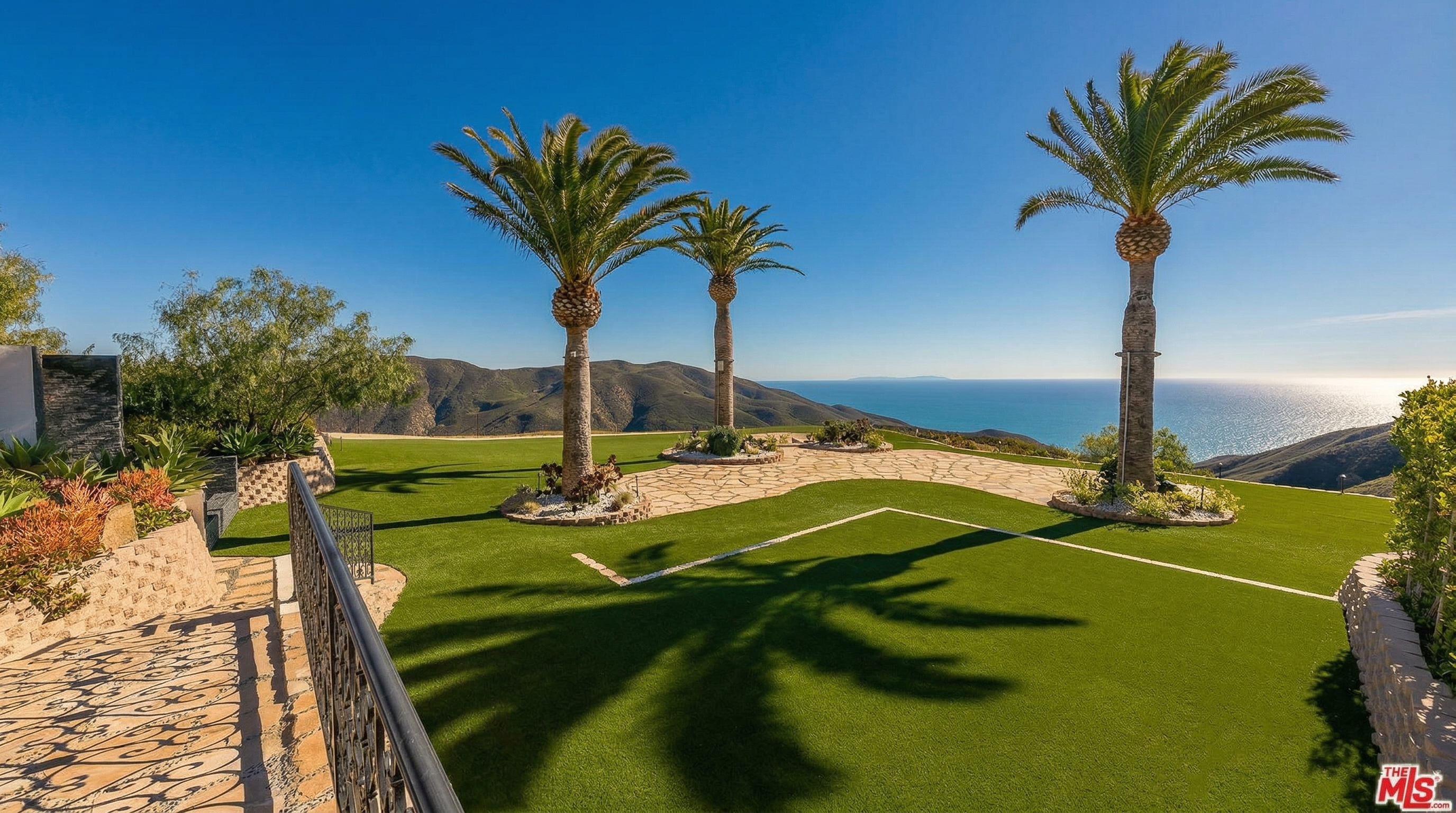 This expansive backyard features a pristine artificial turf lawn framed by mature palm trees and stone-paved pathways. The elevated vantage point offers a breathtaking, unobstructed panoramic view of the ocean and distant coastline under a clear blue sky. The scene conveys a luxurious, serene atmosphere perfect for outdoor entertaining and relaxation.