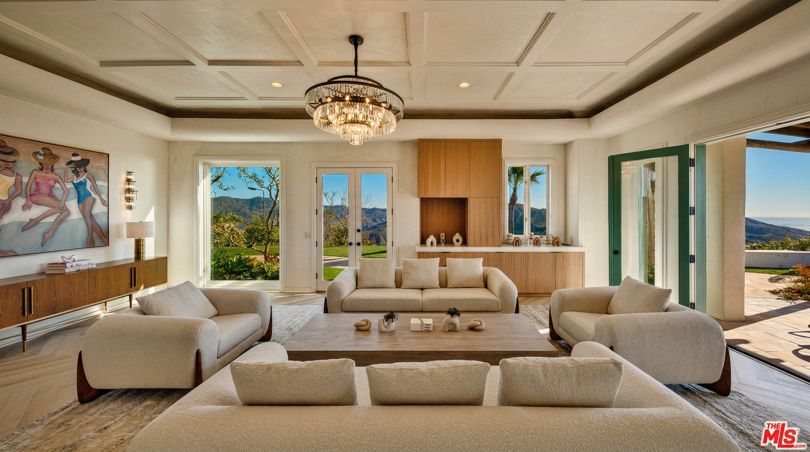 This expansive and bright living room features a sophisticated, neutral color palette with plush, cream-colored sofas arranged around a large, low-profile coffee table. The room is anchored by a stunning coffered ceiling and a grand crystal chandelier, while large windows and glass doors frame breathtaking mountain views. The space exudes a luxurious, airy atmosphere, blending modern elegance with a seamless connection to the outdoors.