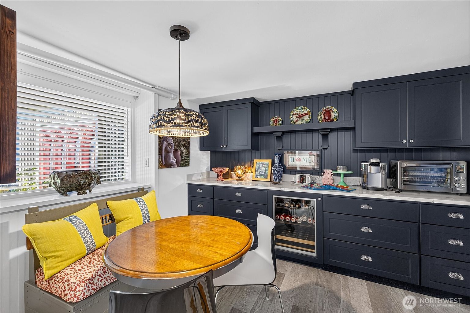 This cozy kitchen area features dark navy cabinetry with a beadboard backsplash and a light-colored countertop. A round wooden dining table is positioned in front of a window with blinds, accompanied by a built-in bench with yellow cushions and a modern white chair. The space includes a built-in wine cooler, a toaster oven, and a decorative pendant light, creating a warm and inviting atmosphere.