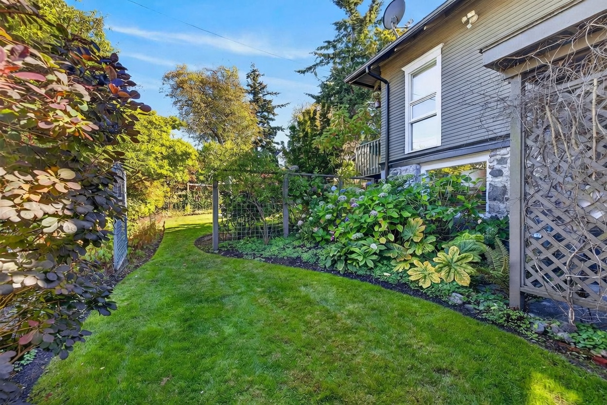 This image captures a lush, well-maintained side yard featuring a vibrant garden bed filled with hydrangeas and various shrubs nestled against the side of a house. The home features dark horizontal siding and a stone-clad foundation, with a wooden trellis providing a decorative element along the garden edge. A manicured lawn curves through the space, leading toward mature trees in the background under a clear blue sky.