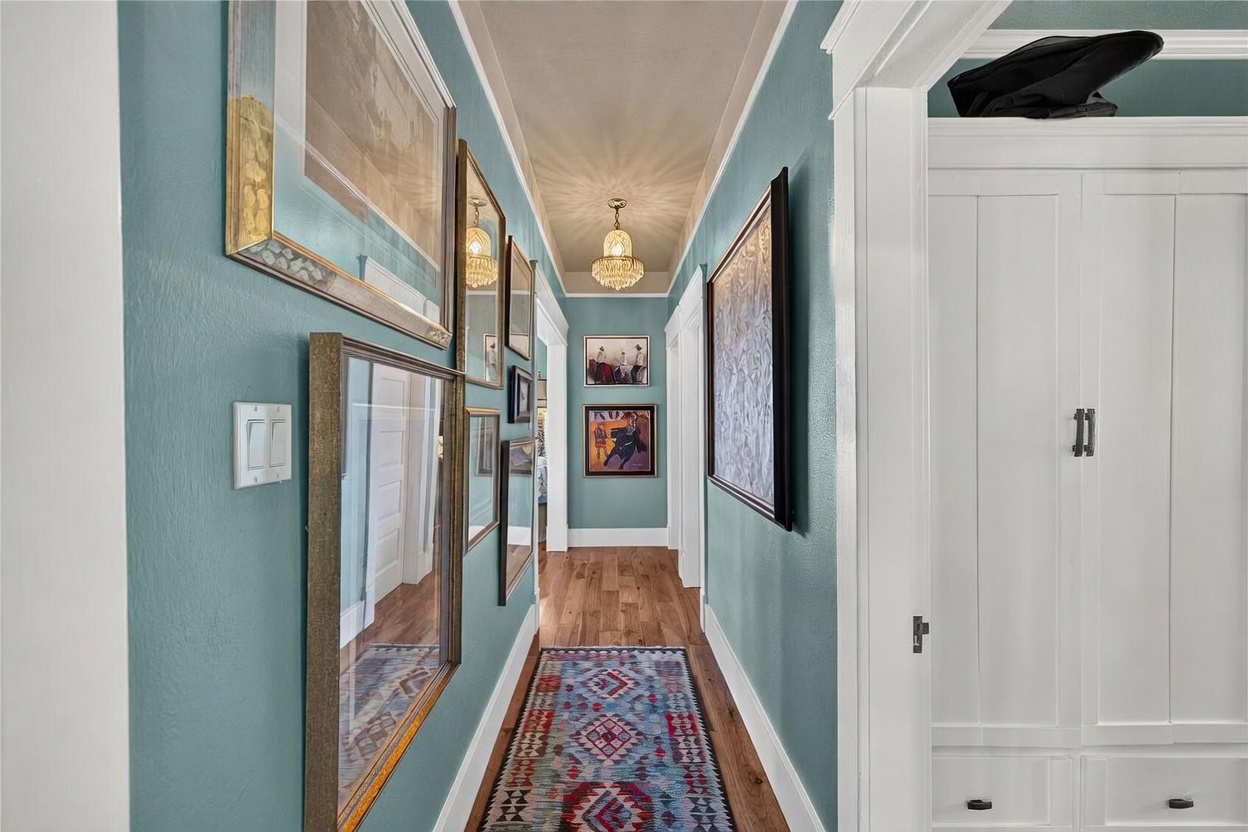This hallway features vibrant teal walls adorned with a gallery of framed artwork and mirrors, creating a sophisticated and personalized aesthetic. The space is anchored by a colorful, patterned runner rug over warm-toned wood flooring and is illuminated by a classic crystal chandelier. A white built-in cabinet on the right provides storage, while the perspective draws the eye down the length of the corridor toward the back of the home.