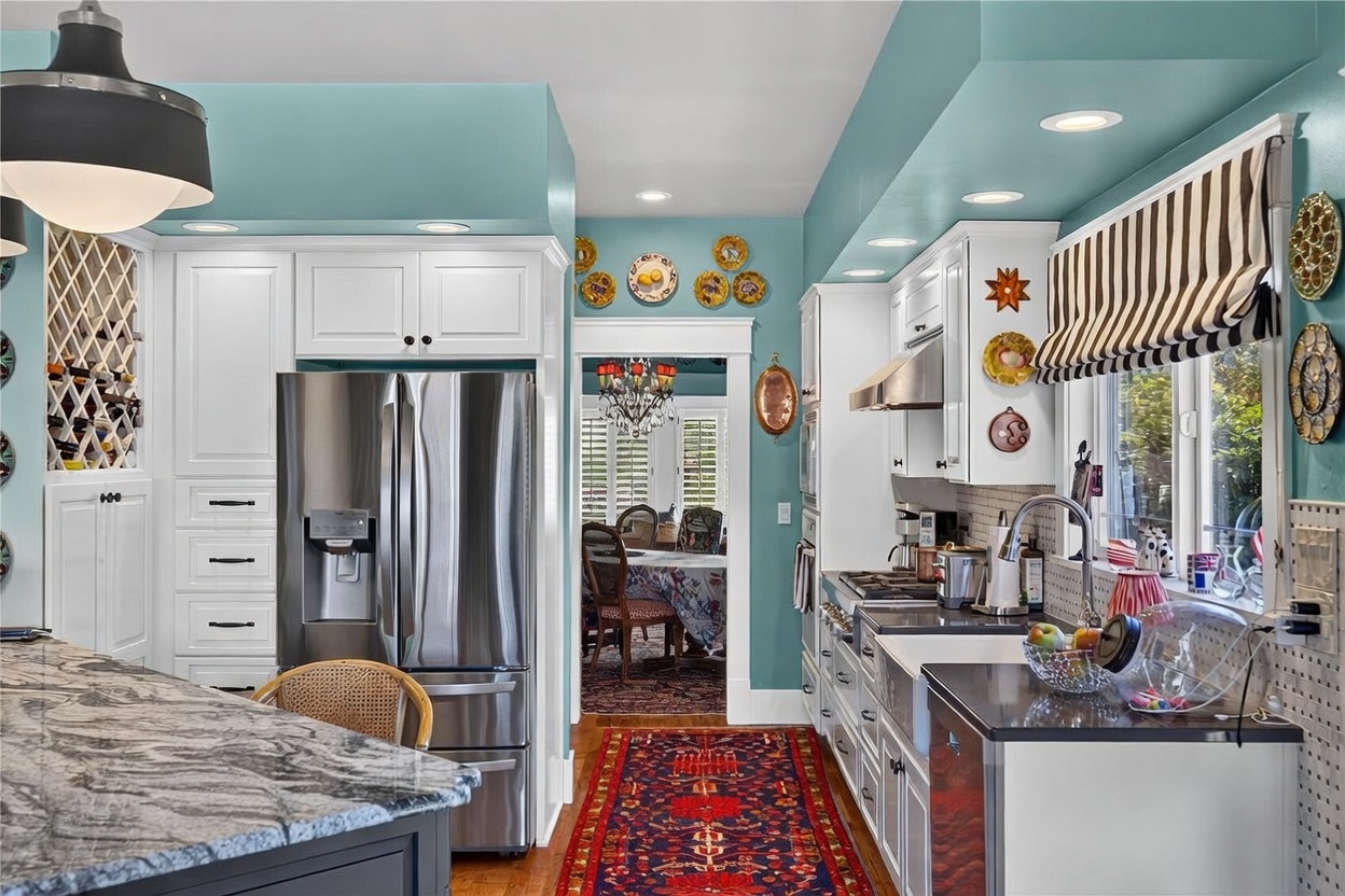 This vibrant kitchen features a striking combination of teal walls, white cabinetry, and dark stone countertops. The space is equipped with stainless steel appliances, including a large refrigerator and a farmhouse-style sink, while decorative plates and a striped window treatment add a personalized, eclectic touch. A view through the doorway reveals a formal dining area, creating a cohesive flow between the two living spaces.