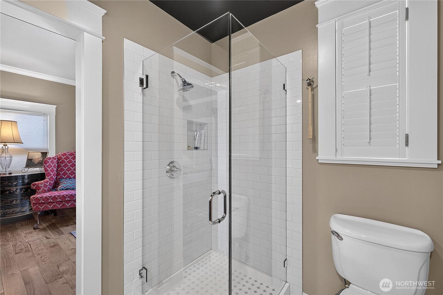 This bathroom features a glass-enclosed shower with white subway tile walls and a built-in recessed niche. To the right, a white toilet sits beneath a window with white plantation shutters, while a doorway on the left provides a glimpse into an adjacent room with a patterned armchair. The space is finished with neutral-toned walls and a dark ceiling, creating a clean and functional aesthetic.