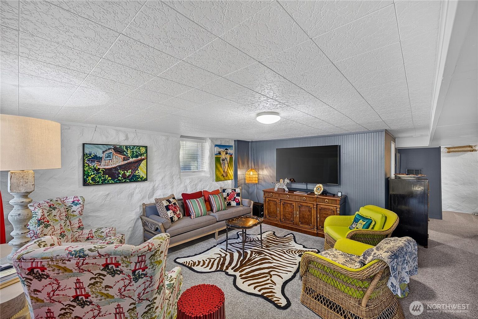 This cozy, eclectic living room features a unique blend of textures, including a white stone-textured wall and blue paneled accent wall. The space is furnished with patterned armchairs, a grey sofa with colorful throw pillows, and two lime green wicker chairs arranged around a zebra-print area rug. A wooden media console sits beneath a wall-mounted television, creating a comfortable and inviting entertainment area.
