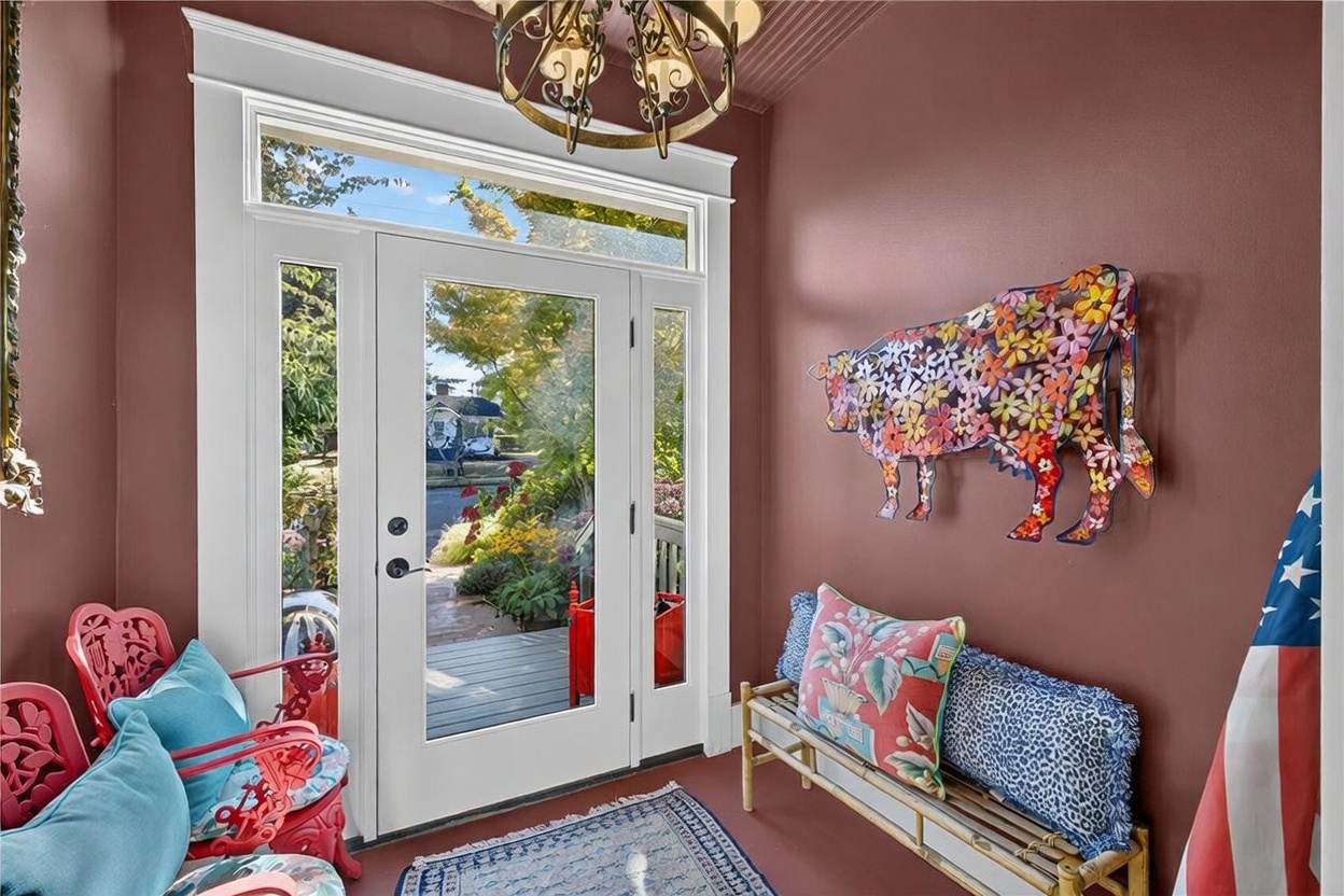 This inviting covered entryway features a warm, deep-toned wall color and a glass-paneled front door that allows natural light to flood the space. The area is furnished with a bamboo-style bench, decorative seating, and a whimsical floral-patterned metal cow wall sculpture, creating a charming and eclectic aesthetic. A patterned area rug anchors the space, while a classic chandelier hangs above, providing a welcoming transition from the outdoors.