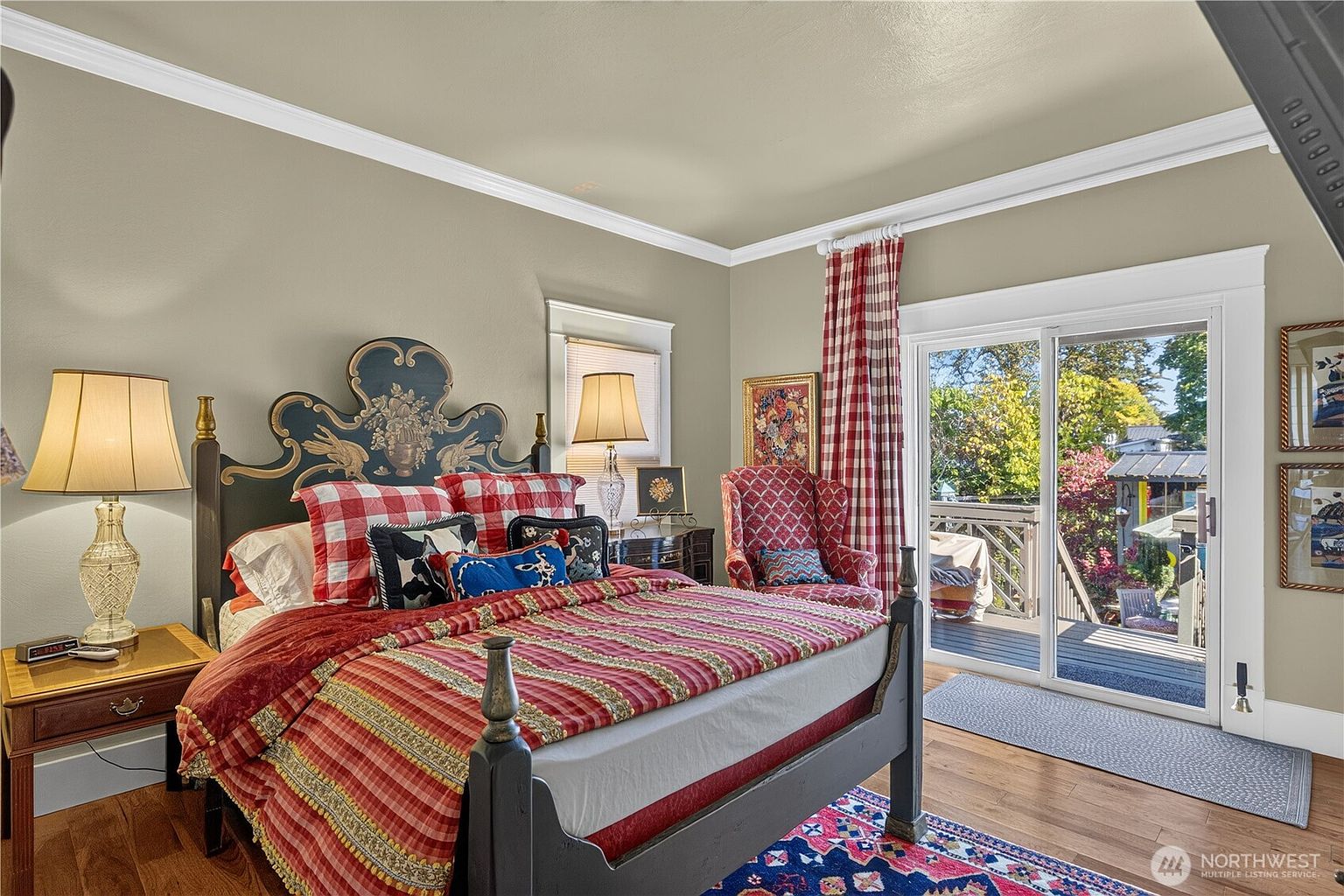This cozy bedroom features a prominent, ornately carved dark-toned headboard and a bed dressed in vibrant red and white plaid bedding. A matching armchair sits near a sliding glass door that opens onto an outdoor deck, allowing for natural light to fill the space. The room is decorated with a patterned area rug, traditional table lamps, and framed artwork, creating a warm and eclectic atmosphere.