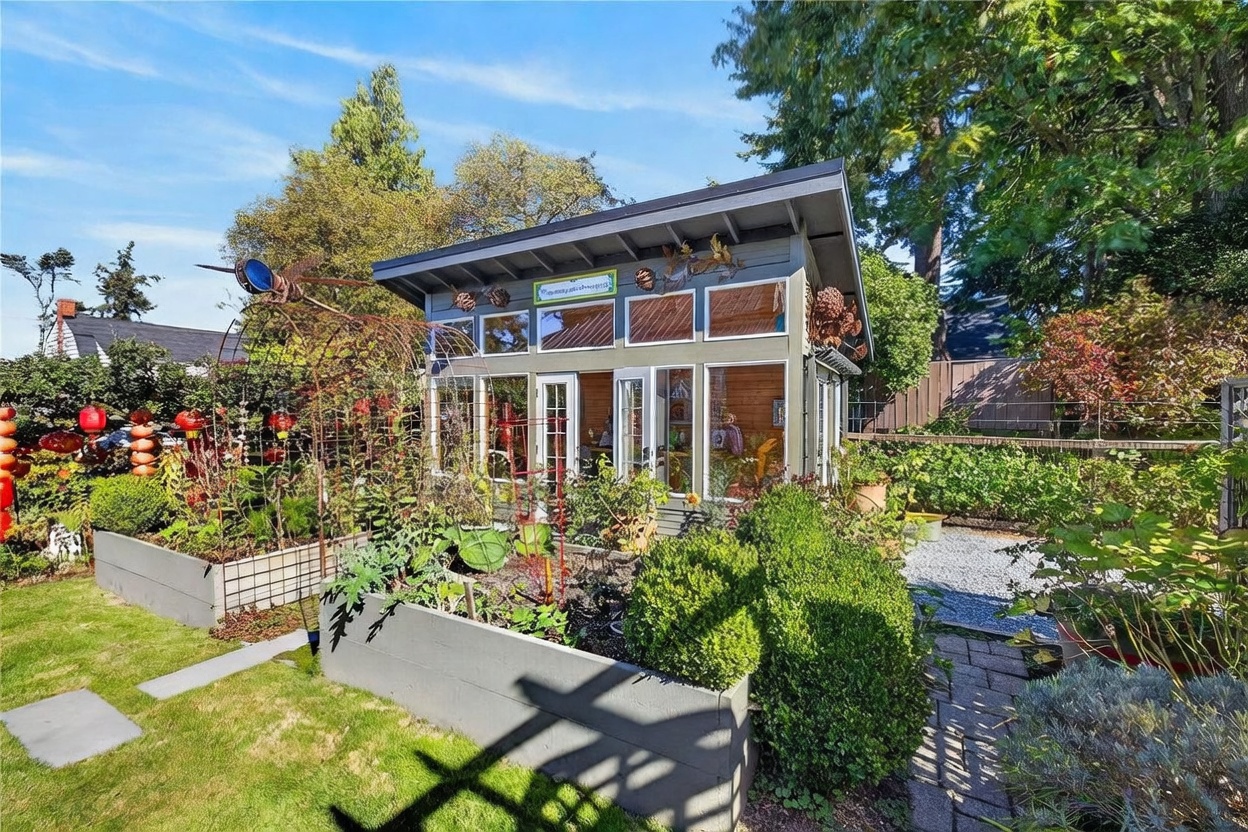 This charming backyard garden features a modern, shed-style studio with large windows and a light-colored wood interior visible through the glass. The structure is surrounded by raised garden beds, a metal trellis, and lush greenery, creating a serene and functional outdoor retreat. The scene is captured from a slightly elevated perspective, highlighting the well-maintained landscaping and the inviting, sunlit atmosphere of the garden space.