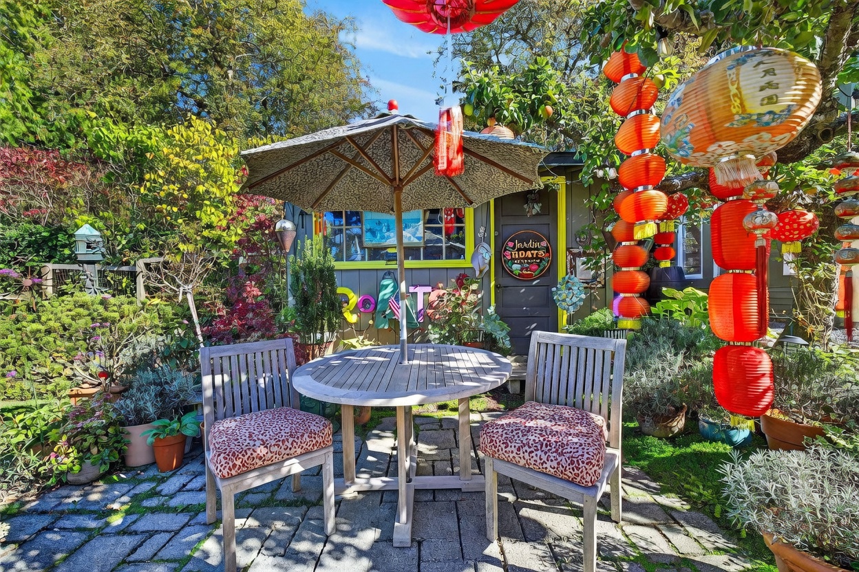This charming backyard garden space features a stone-paved patio area centered around a round wooden table with two matching chairs topped with patterned cushions. A small, rustic shed with a bright yellow door frame serves as a focal point, adorned with decorative signage and surrounded by lush greenery and vibrant hanging paper lanterns. The scene is captured from a low, eye-level perspective, emphasizing the cozy, whimsical atmosphere of this private outdoor retreat.