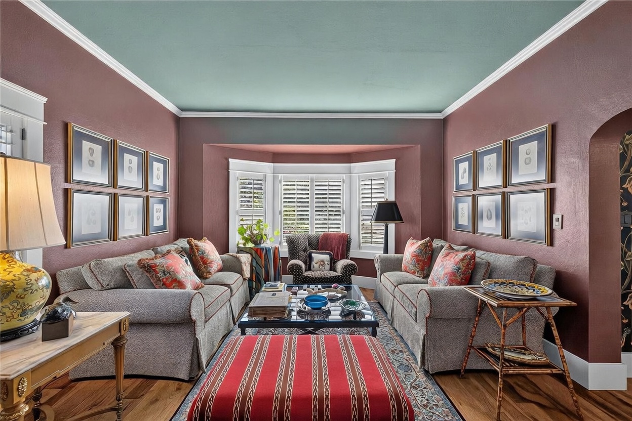 This cozy living room features deep mauve walls contrasted by a soft teal ceiling and white crown molding. The space is anchored by two matching sofas facing each other, a central glass-topped coffee table, and a large red-striped ottoman, all set upon a patterned area rug. A bay window with white shutters serves as the focal point, flanked by symmetrical wall art and a comfortable armchair.