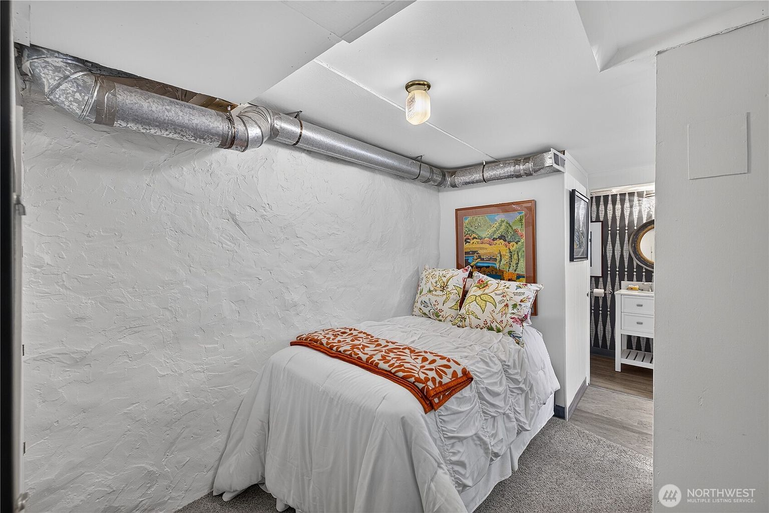 This cozy bedroom is located in a basement setting, characterized by a textured white stone wall and exposed overhead ductwork. The room features a single bed with a white comforter and patterned pillows, positioned beneath a framed landscape print. A doorway leads into an adjacent bathroom with a vanity and patterned wallpaper, creating a compact and functional living space.