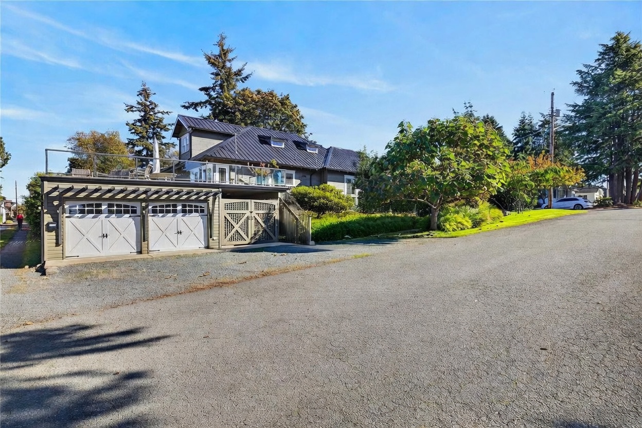 This exterior view captures a charming home with a prominent detached garage featuring a rooftop deck enclosed by glass railings. The house is set on a gentle slope, surrounded by mature trees and a well-maintained lawn, with a paved street leading up to the property. The architectural style blends traditional elements with modern outdoor living spaces, creating an inviting and well-integrated curb appeal.