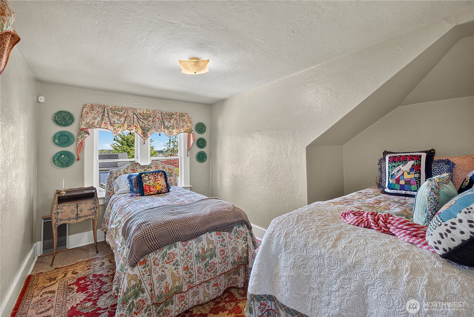 This cozy bedroom features two twin beds with patterned bedding and decorative pillows, set against a sloped ceiling characteristic of an attic-style room. A small wooden side table sits near a window adorned with a floral valance, while decorative plates are mounted on the wall. The room has a warm, traditional aesthetic with a patterned area rug covering the floor.