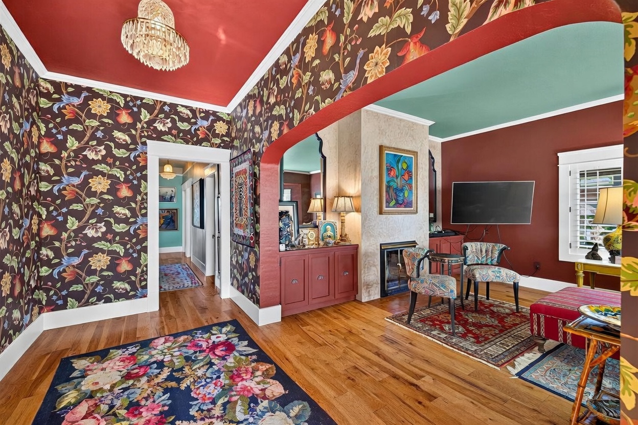 This eclectic living space features bold, dark floral wallpaper and a vibrant red ceiling, creating a dramatic and highly personalized aesthetic. The room includes a tall grandfather clock, a fireplace with a textured surround, and a seating area arranged on a patterned rug. The perspective looks from a hallway entrance into the living area, showcasing the open transition between the two spaces.