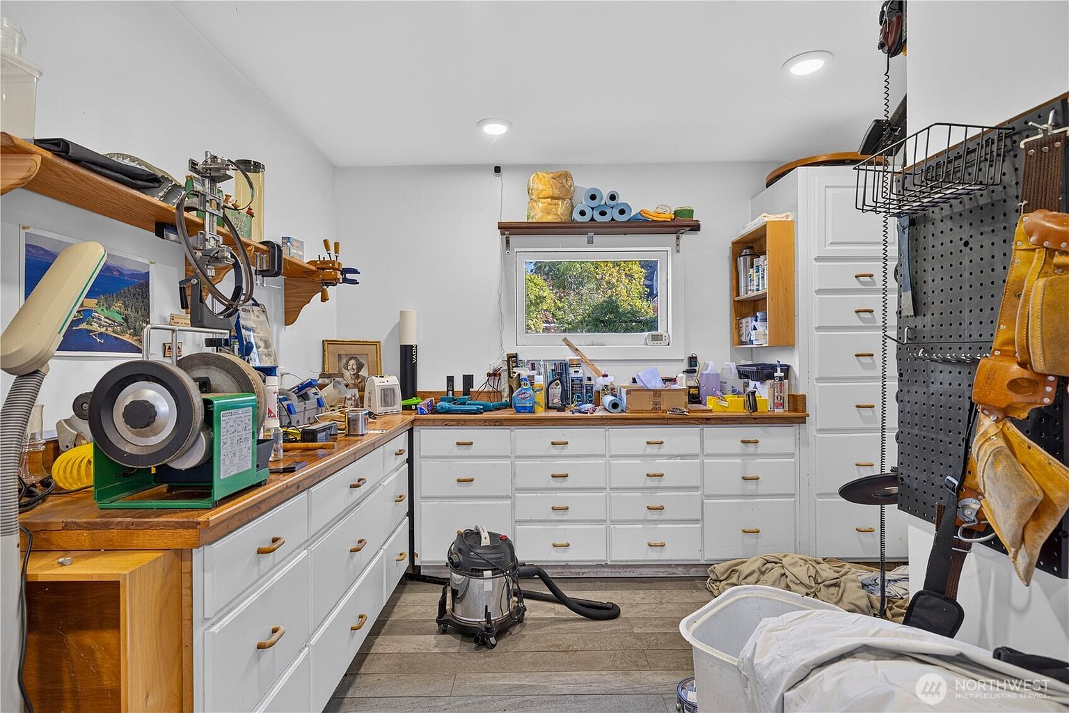 This image depicts a functional workshop or hobby room featuring extensive white cabinetry and a long, sturdy wooden workbench. The space is equipped with various tools, including a bench grinder, and includes a pegboard wall for organized storage, making it an ideal area for DIY projects or crafts. A central window provides natural light, while the room's layout emphasizes utility and practical storage solutions.