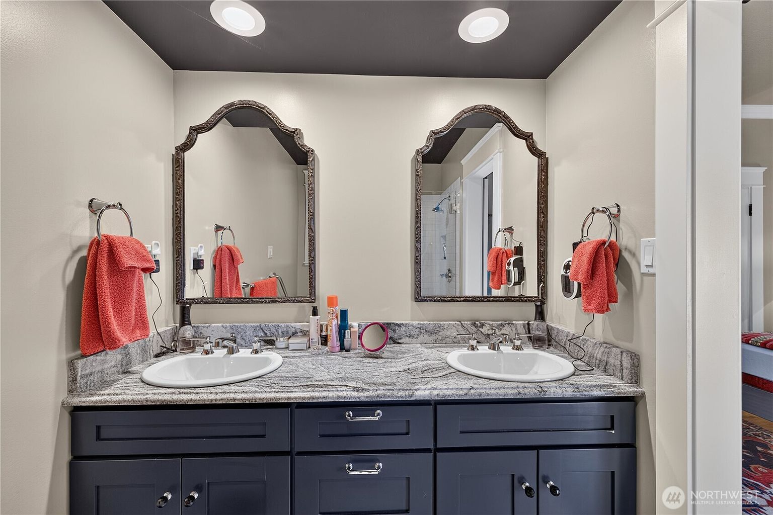 This primary bathroom features a double vanity with dark cabinetry topped by a stone countertop and two oval undermount sinks. Above the vanity, two ornate, dark-framed mirrors are mounted on a light-colored wall, complemented by recessed lighting in the dark-painted ceiling. The space is accented with bright orange towels and personal grooming items, creating a functional and well-lit vanity area.