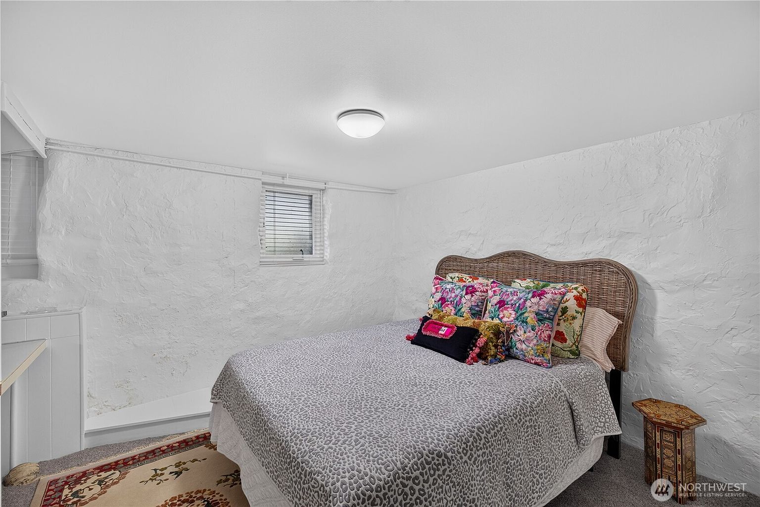 This cozy bedroom features a textured, white-painted stone wall that adds a rustic, cottage-like character to the space. The room is furnished with a bed topped with a patterned grey coverlet and a variety of colorful, floral-print throw pillows, complemented by a woven headboard and a small, decorative side table. A window with horizontal blinds allows natural light to enter, while a simple ceiling light fixture provides overhead illumination.