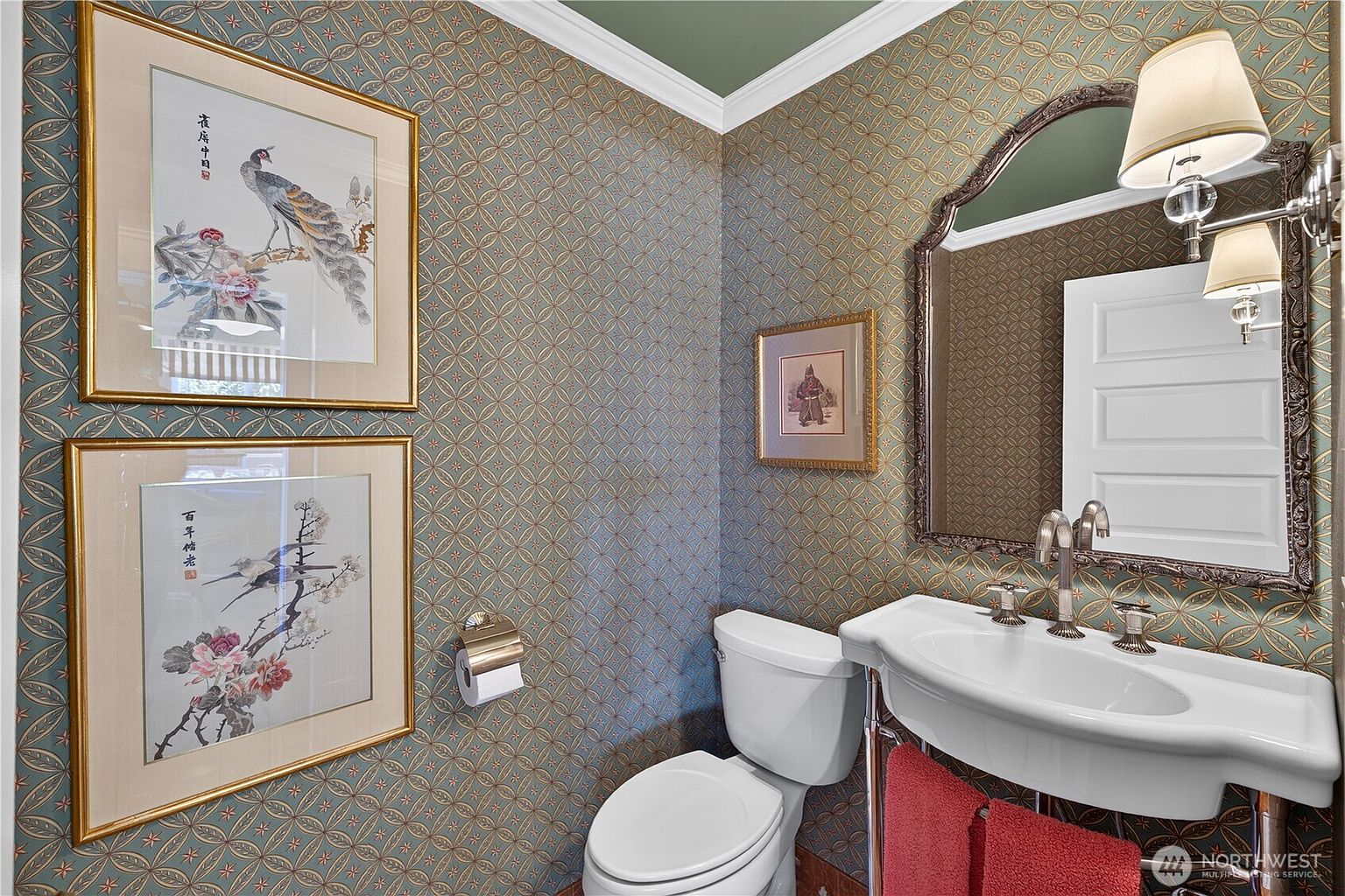 This elegant powder room features intricate patterned wallpaper in shades of blue and gold, complemented by a pedestal sink and a decorative arched mirror. Two framed bird-themed art pieces hang on the wall, adding a sophisticated, classic touch to the space. The room is illuminated by a dual-light wall fixture, creating a warm and inviting atmosphere.